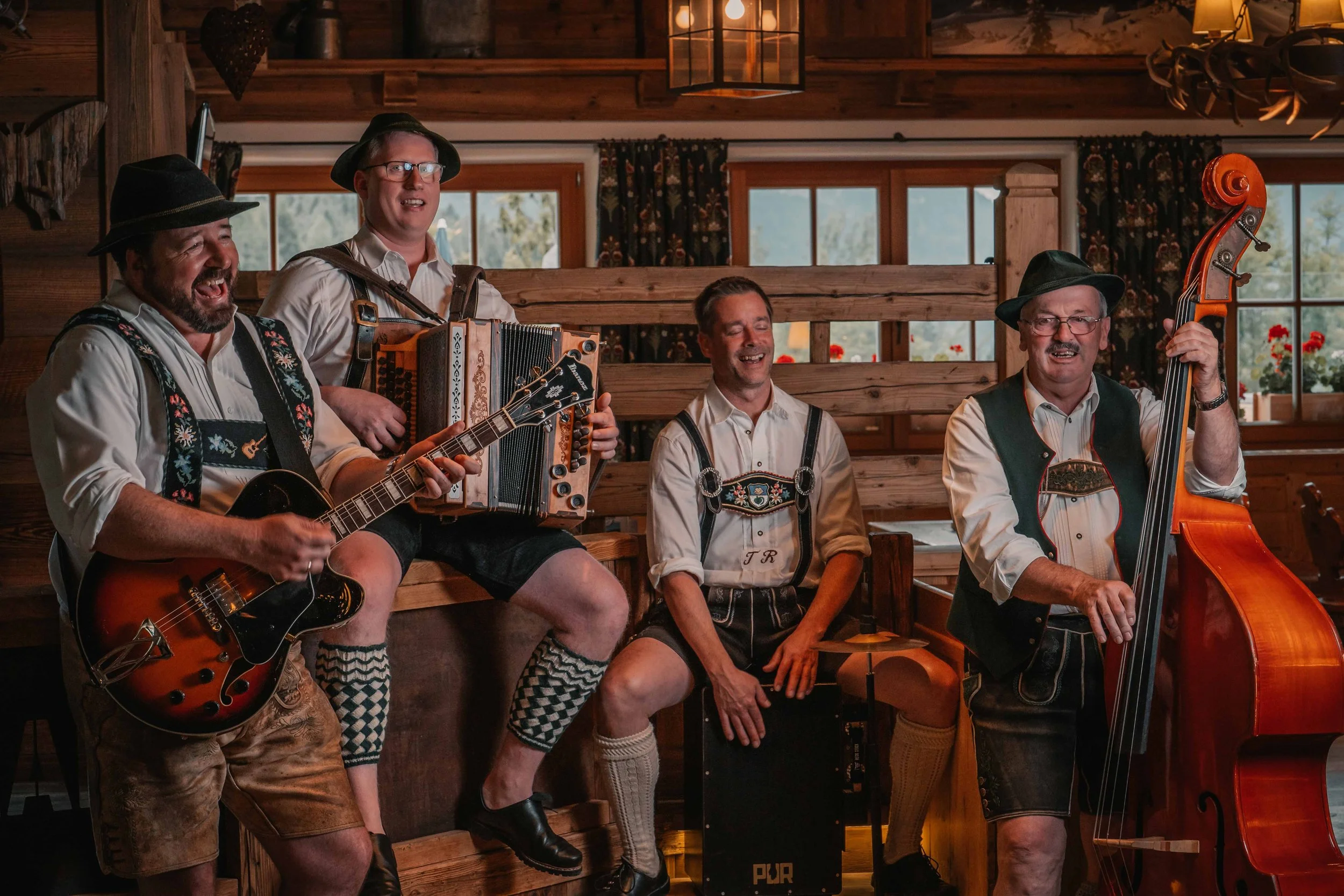 Eine Gruppe von vier Männern in traditioneller bayrischer Kleidung, die in einer Holzhütte Volksmusik spielen und singen, während sie verschiedene Instrumente wie Gitarre, Akkordeon, Cajón und Kontrabass spielen.