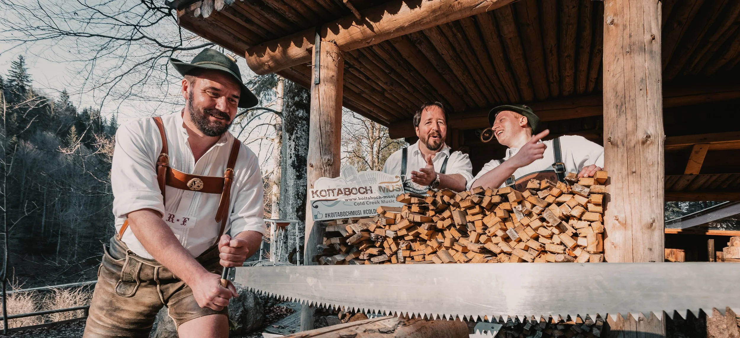 Drei Männer in traditioneller Tracht unterhalten sich in einer Holzhütte, während einer mit einer großen Säge Holz schneidet. Im Hintergrund sind Bäume und ein klarer Himmel sichtbar.