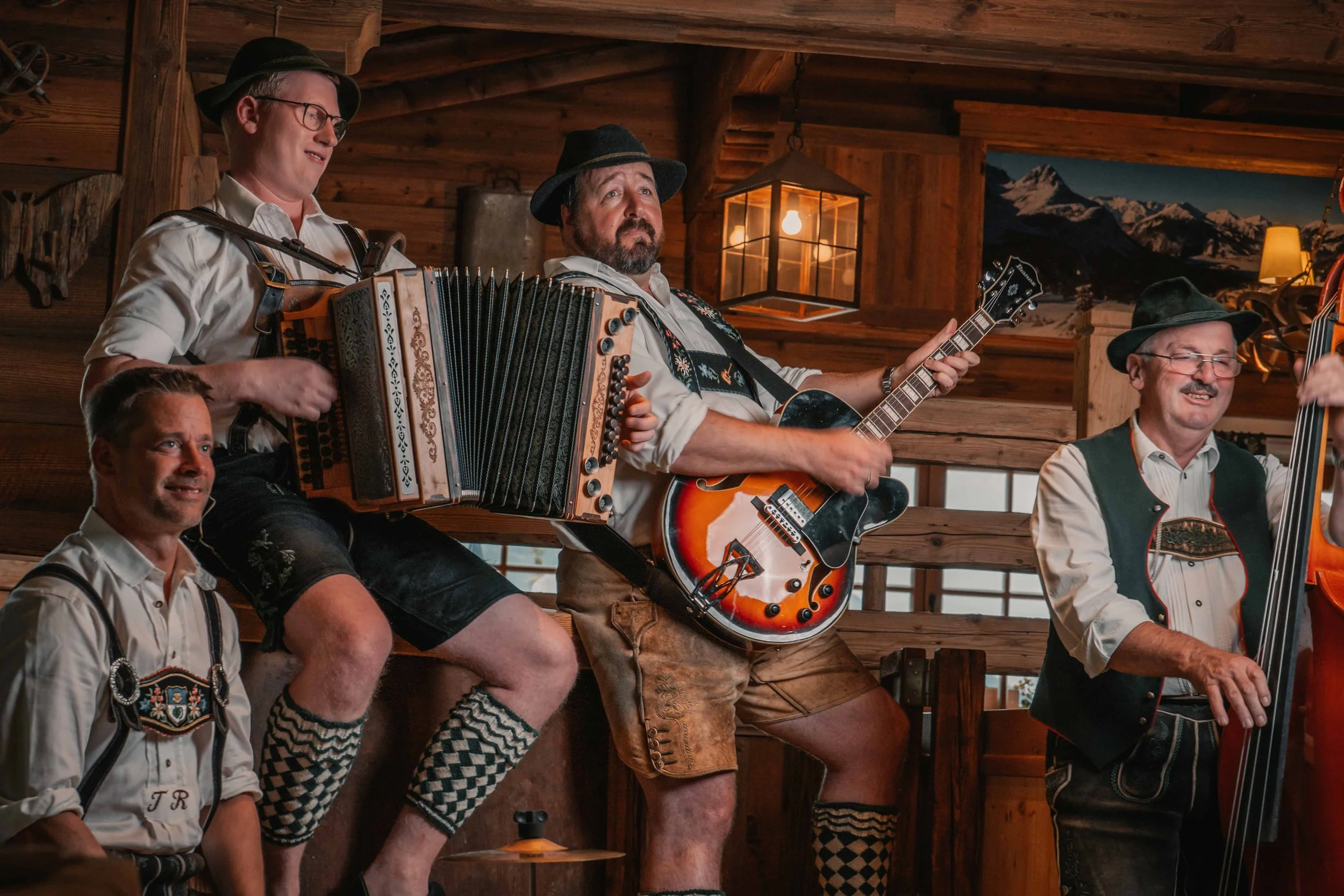 Gruppen von Männern in traditioneller bayerischer Kleidung spielen Musik in einer Holzhütte, einige mit Instrumenten wie Gitarre und Akkordeon.