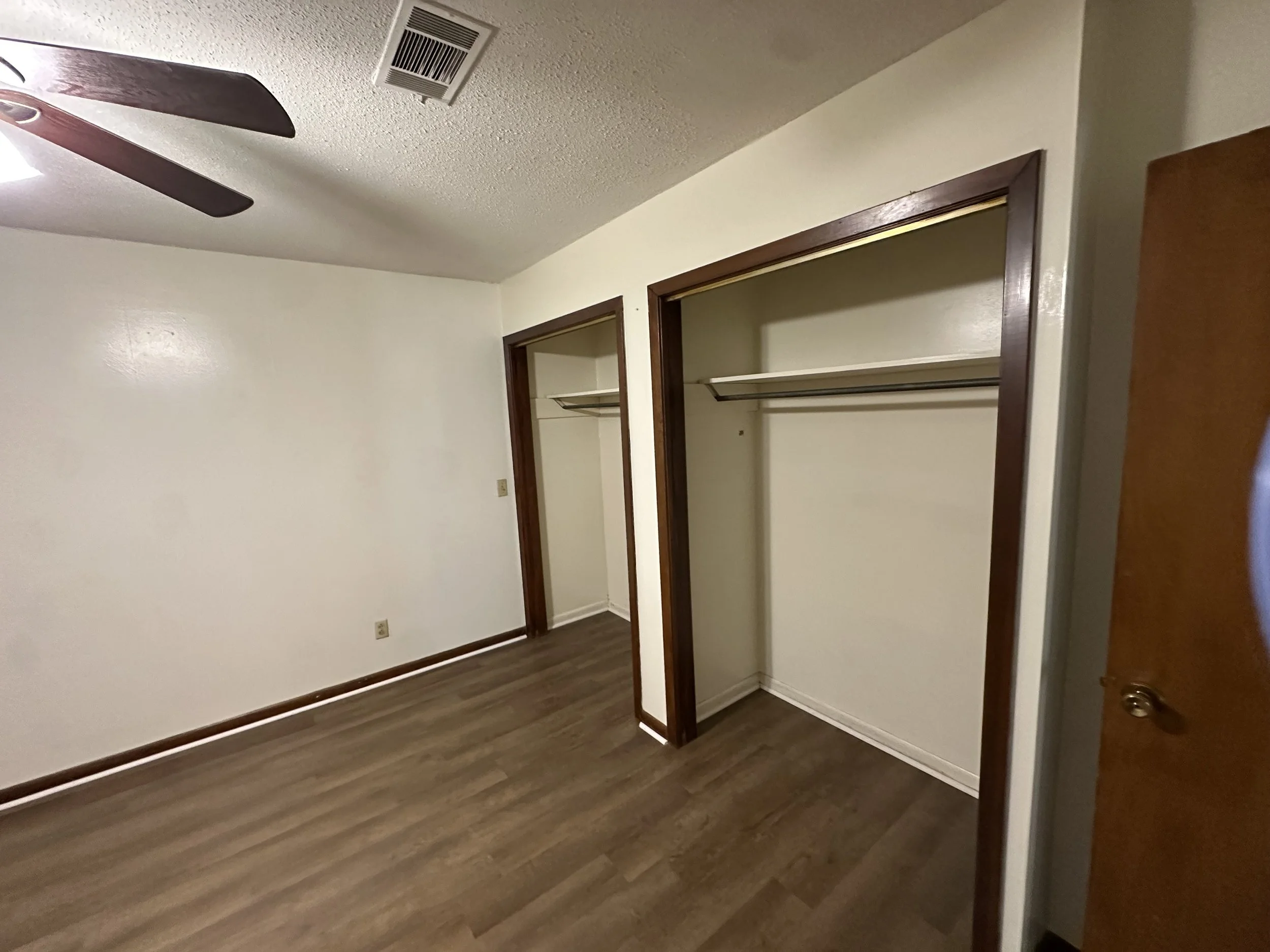 Empty bedroom with two open closet spaces with wood trim, white walls, a ceiling fan, and hardwood flooring.