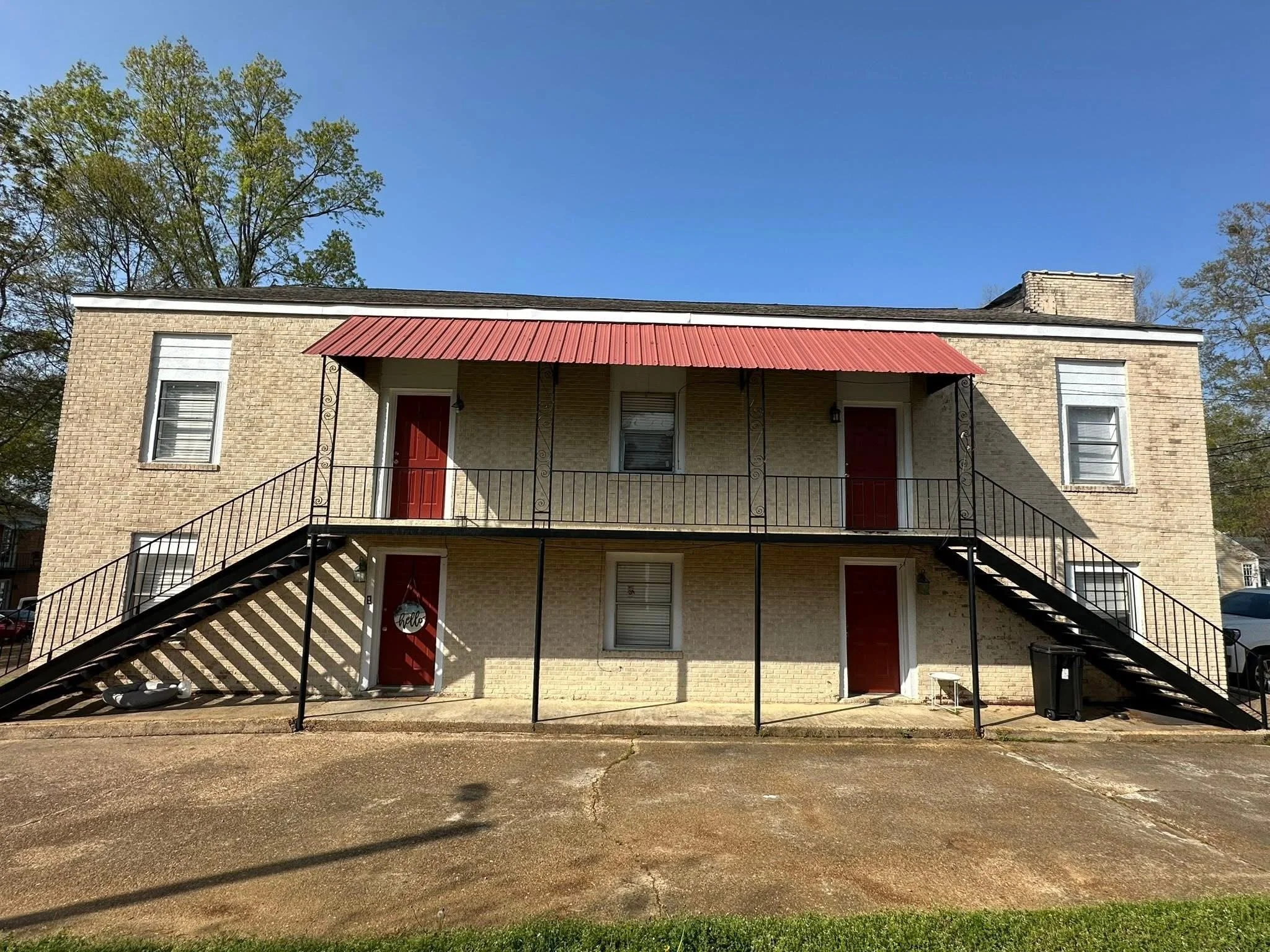 Two-story brick apartment building with four red doors, black metal staircases on each side, and a red metal awning over the upper balcony. The building has white trim around the windows, and a parking lot in front with green grass at the bottom. There are trees and a blue sky in the background.