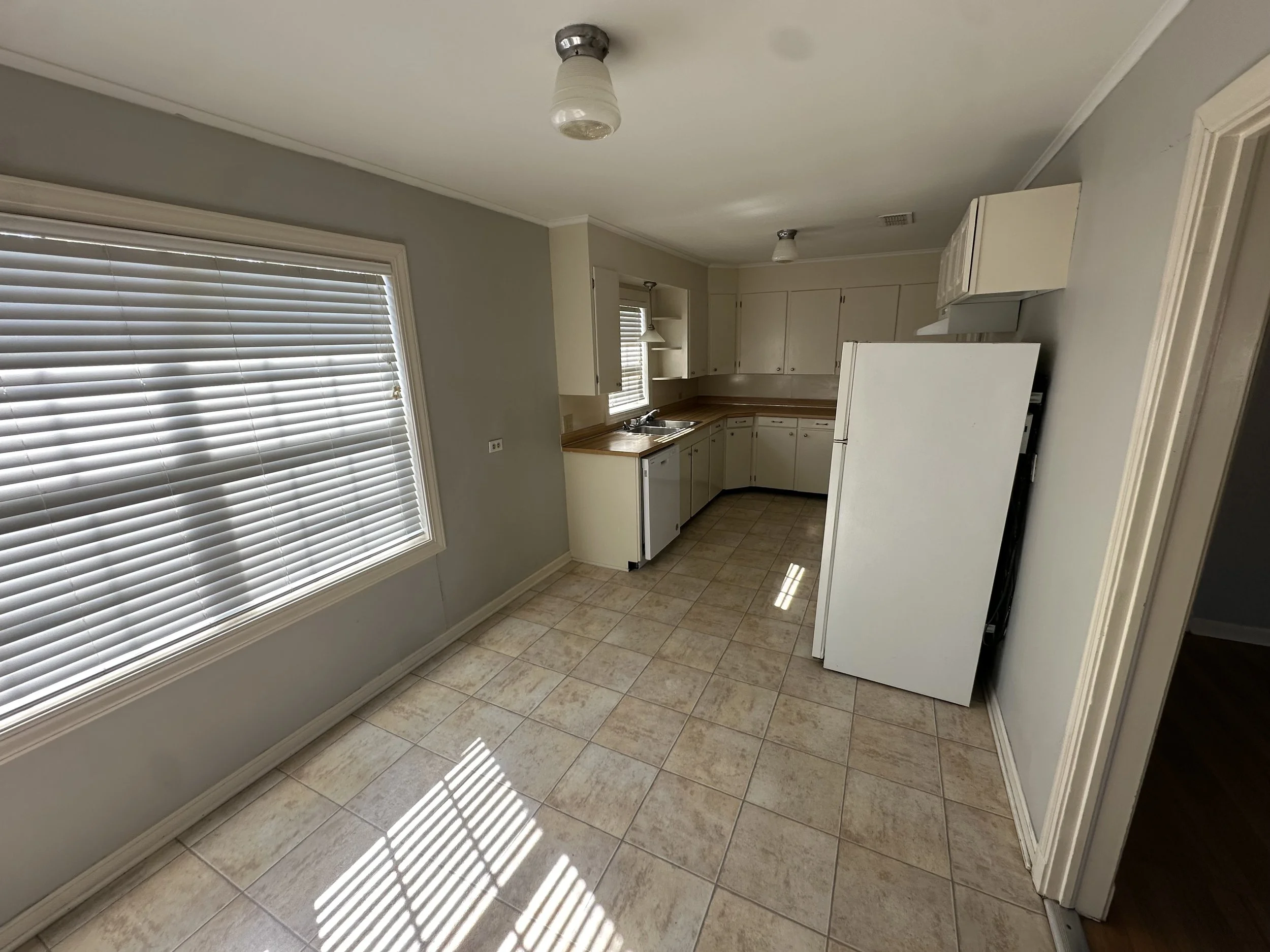 Empty kitchen with large window blinds, beige tiled floor, white cabinets, and a white refrigerator.