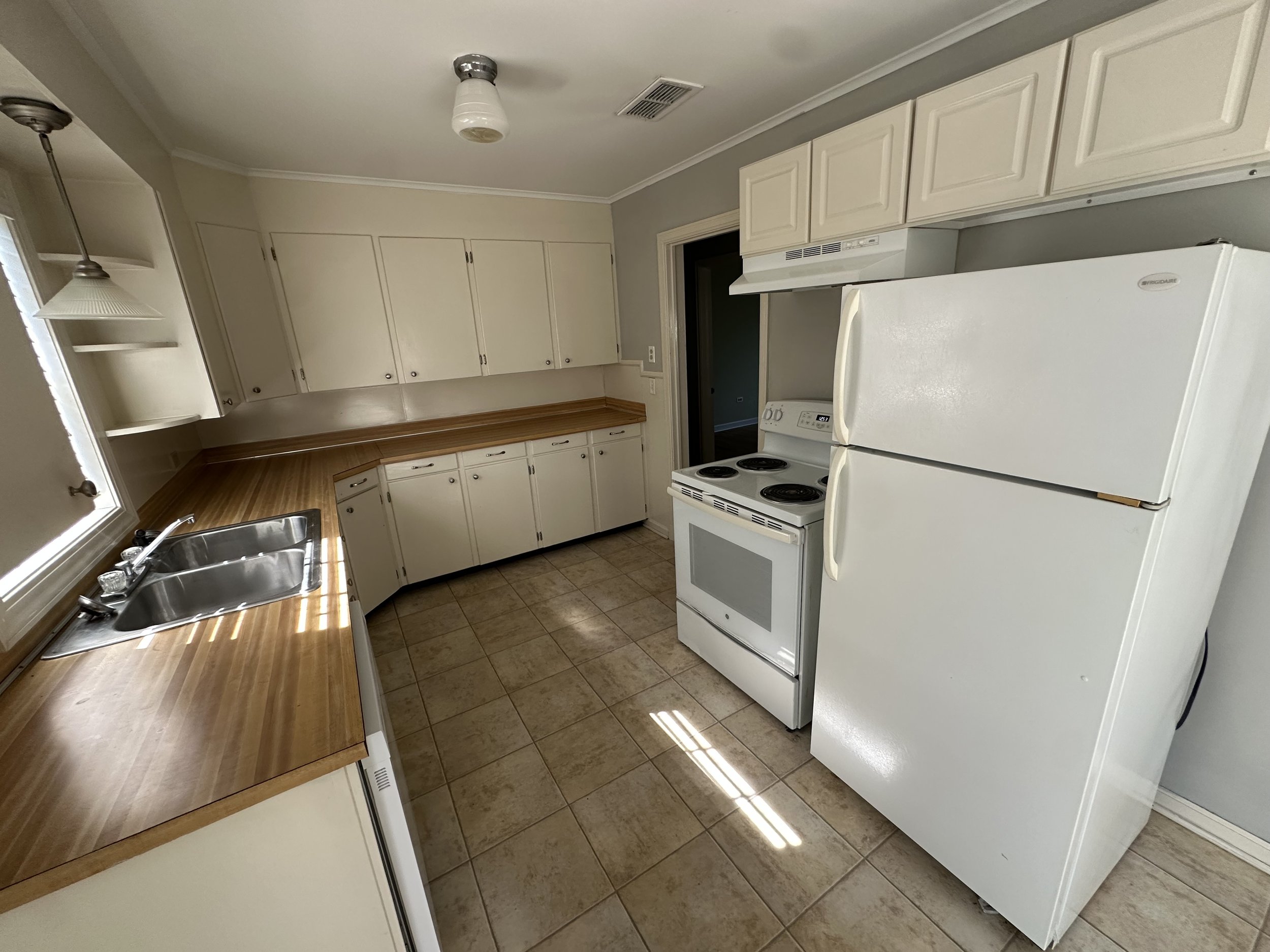 Empty kitchen with white cabinets, wooden countertops, a white refrigerator, white stove, stainless steel sink, tiled floor, window, and empty shelves.