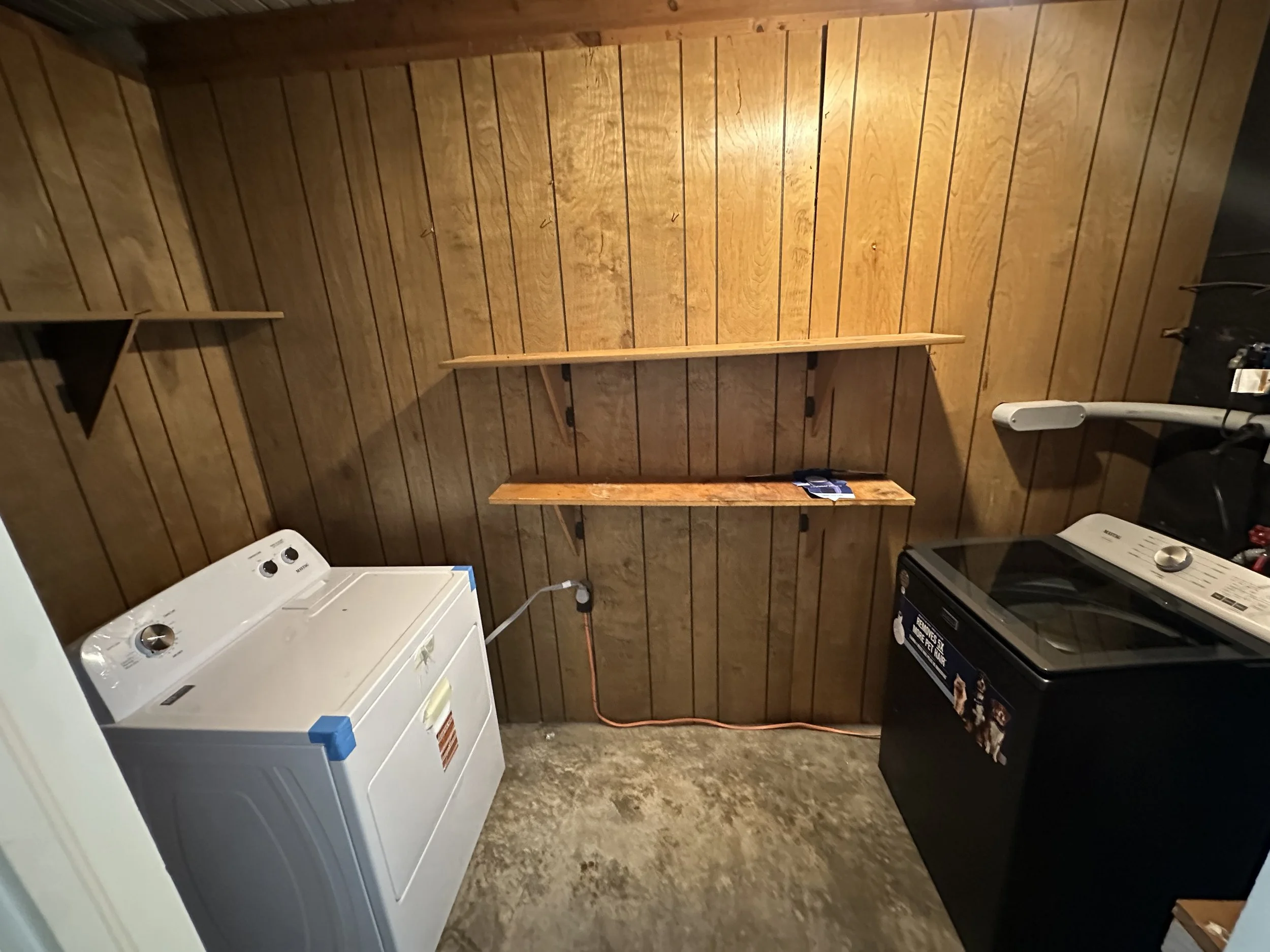 A laundry room with wood-paneled walls, a white top-loading washing machine on the left and a black top-loading dryer on the right, with two wooden shelves on the wall above the appliances, and a concrete floor.