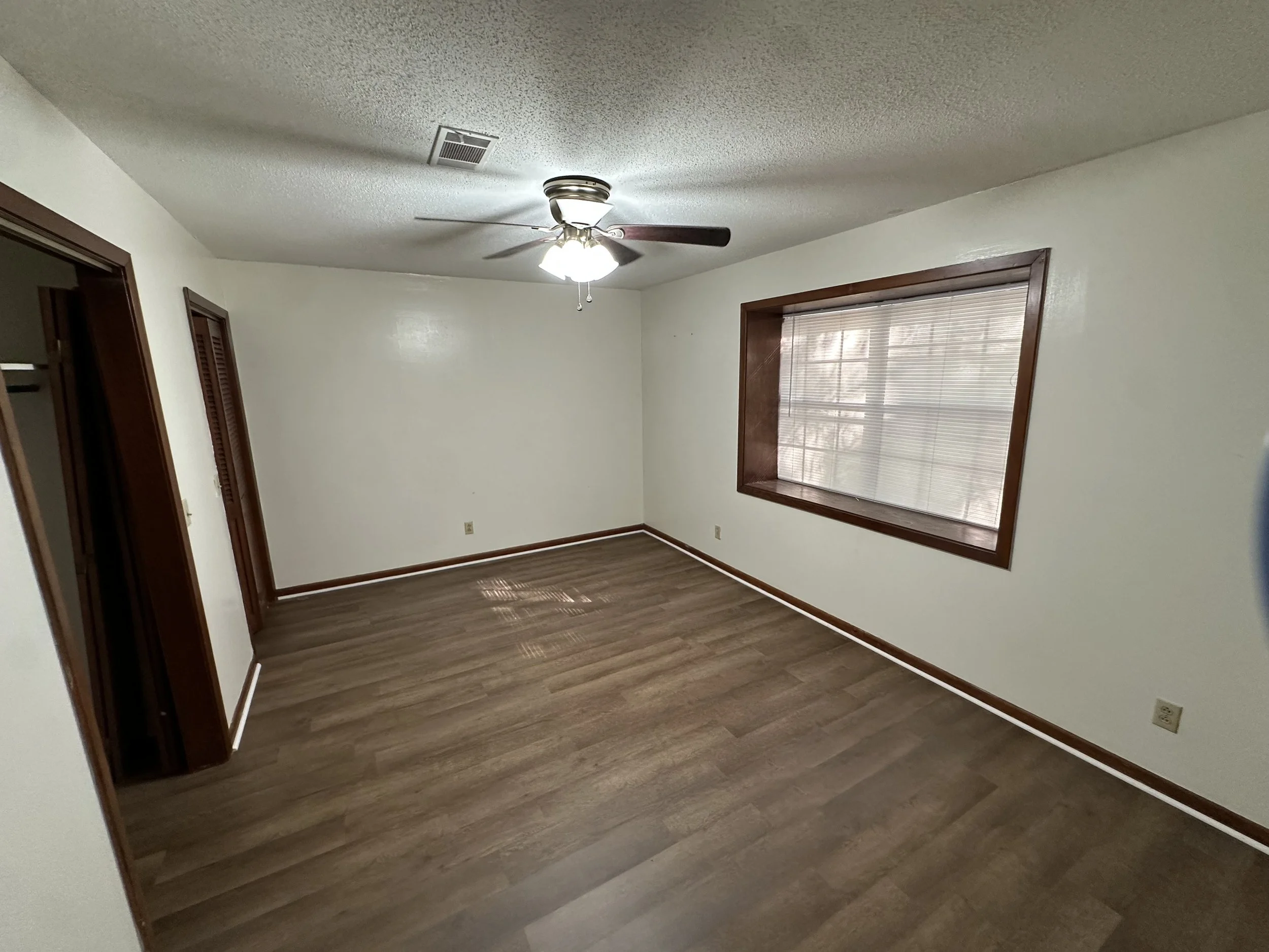 An empty rectangular room with white walls, a ceiling fan with light, a large window with blinds, and wooden trim around the window and doorways. The floor is wood or laminate.
