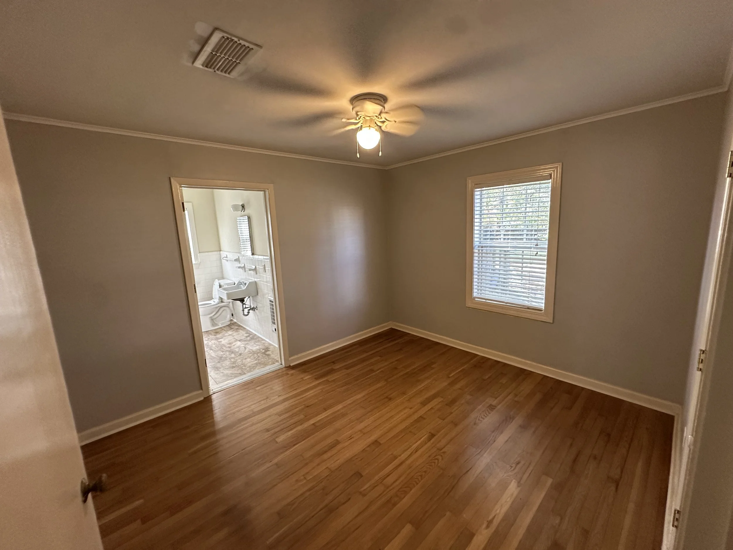 Empty bedroom with hardwood floors, a ceiling fan with light, a window with blinds, and a doorway leading to a small bathroom with a toilet and sink.