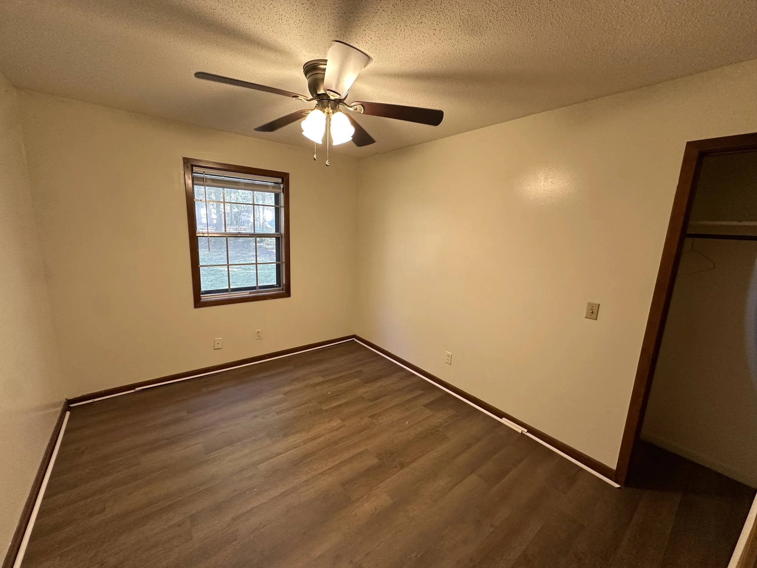 Empty bedroom with a window, ceiling fan, wooden flooring, and an open closet.