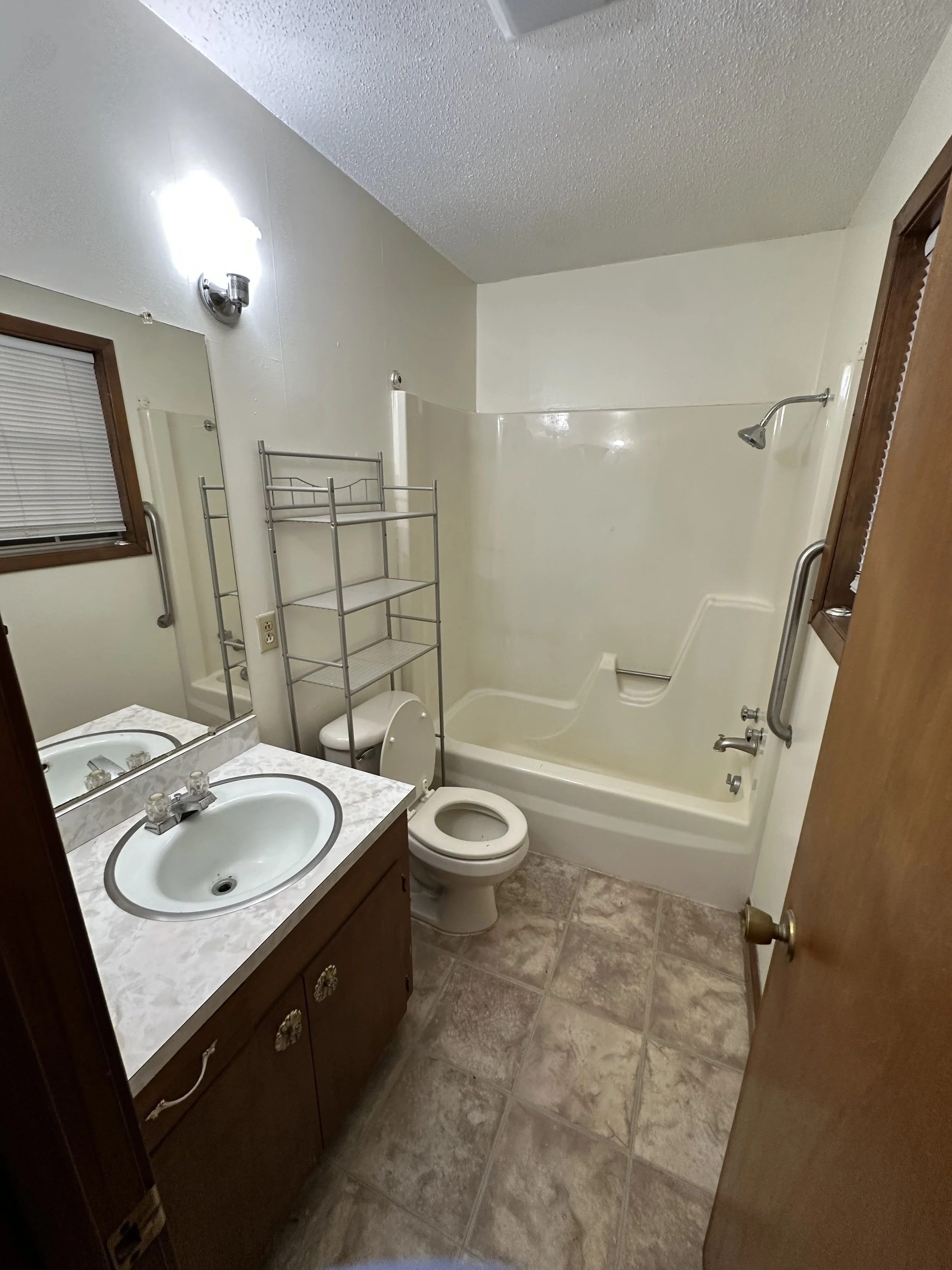 Small bathroom with a toilet, sink with a mirror, bathtub with a shower, window with blinds, shelves above toilet, and beige tile floor.