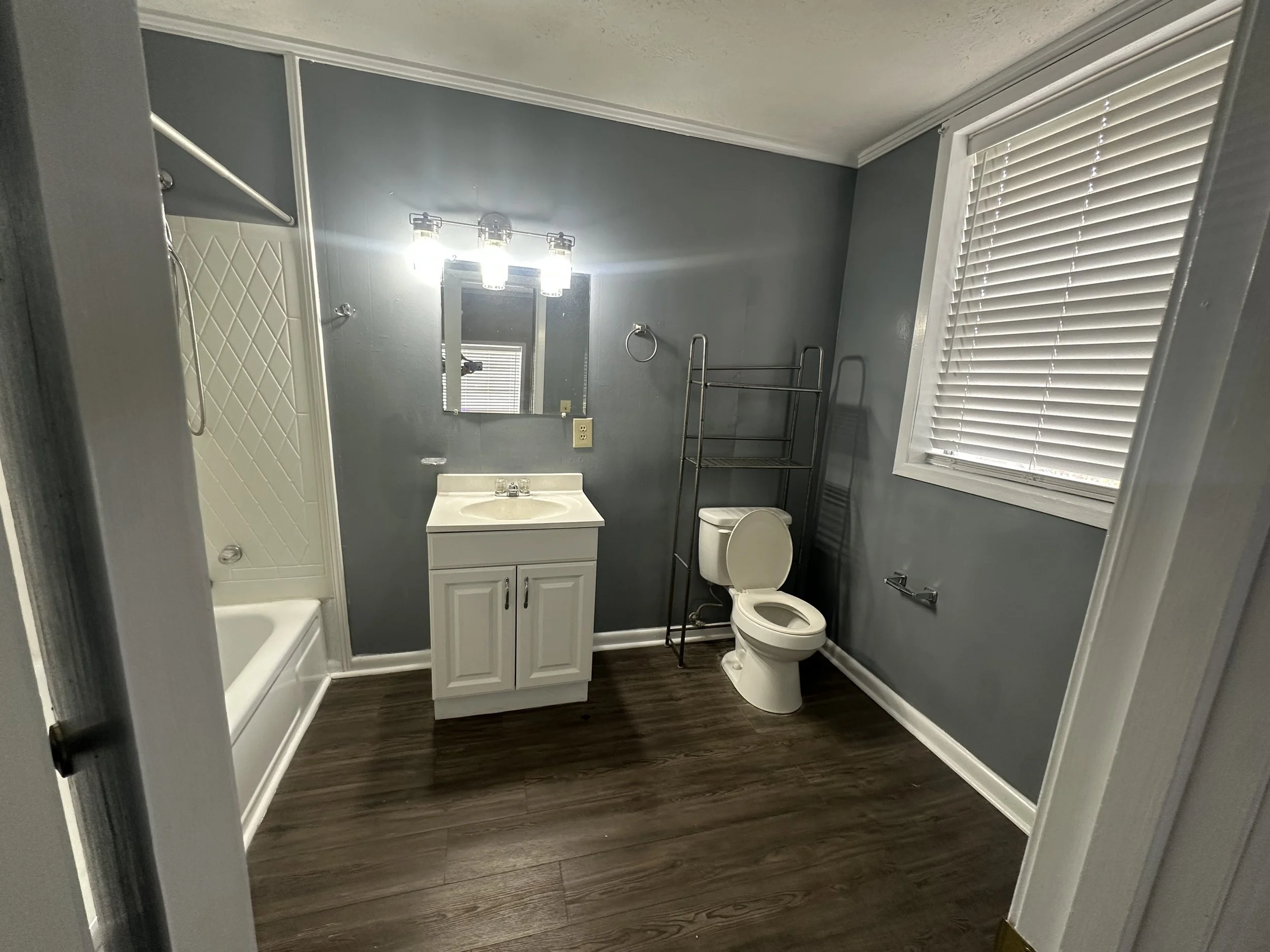 Bathroom with a shower on the left, a white vanity with a mirror and three-light fixture, a toilet, a metal shelving unit, a window with blinds, and dark wood flooring.