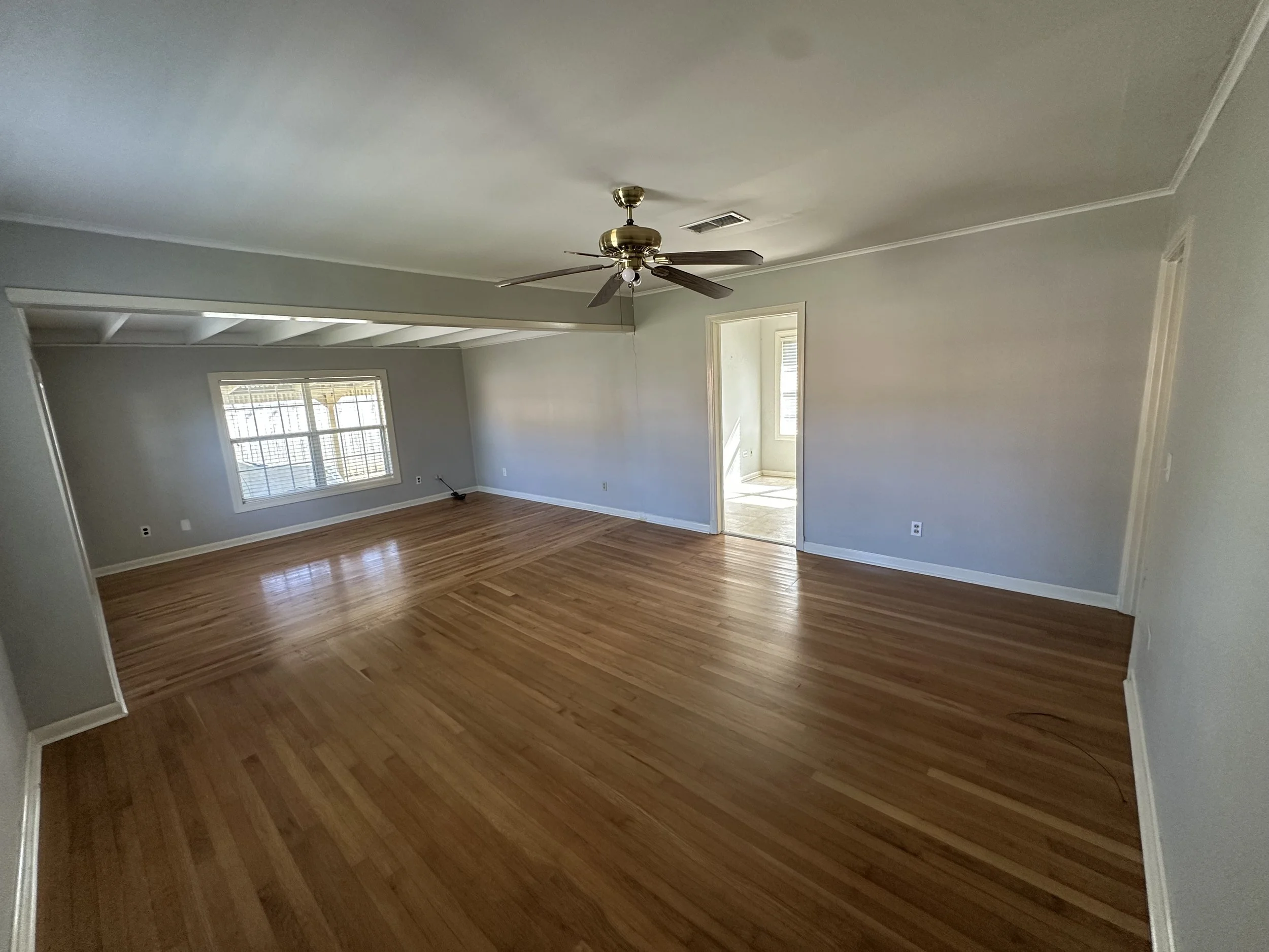 Empty living room with hardwood floors, light gray walls, a ceiling fan, and a window.