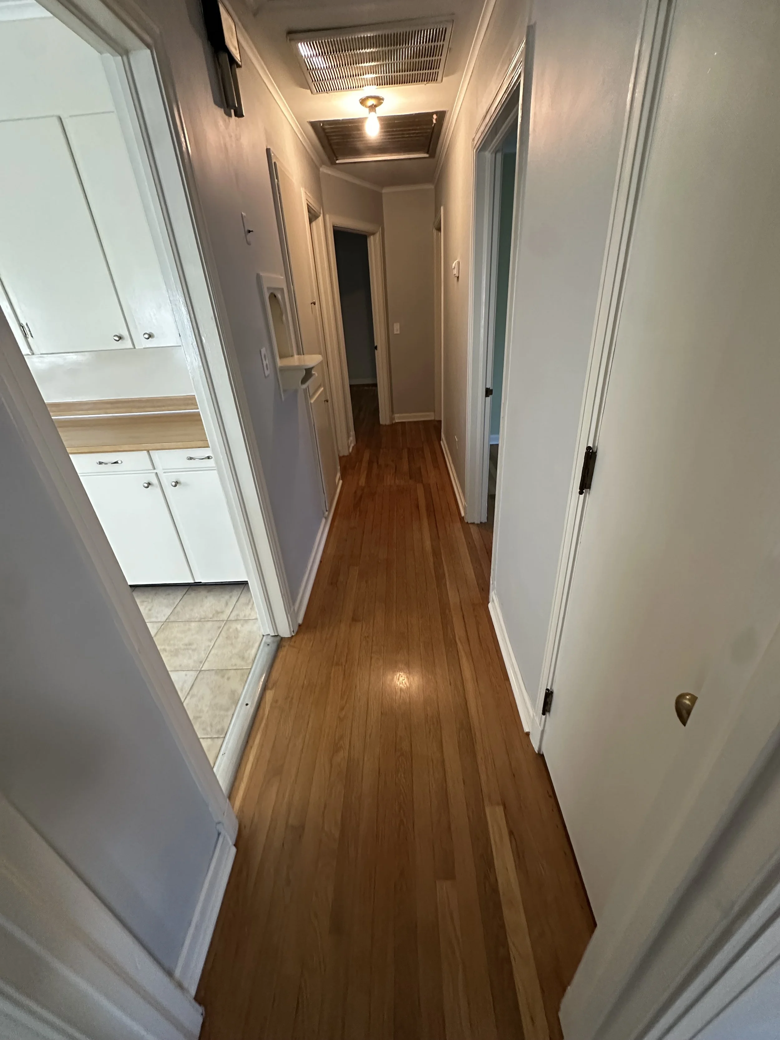 Empty hallway with wooden flooring, white walls, and multiple closed doors. The ceiling has a light fixture and ventilation grilles, with the kitchen visible to the left through an open door.