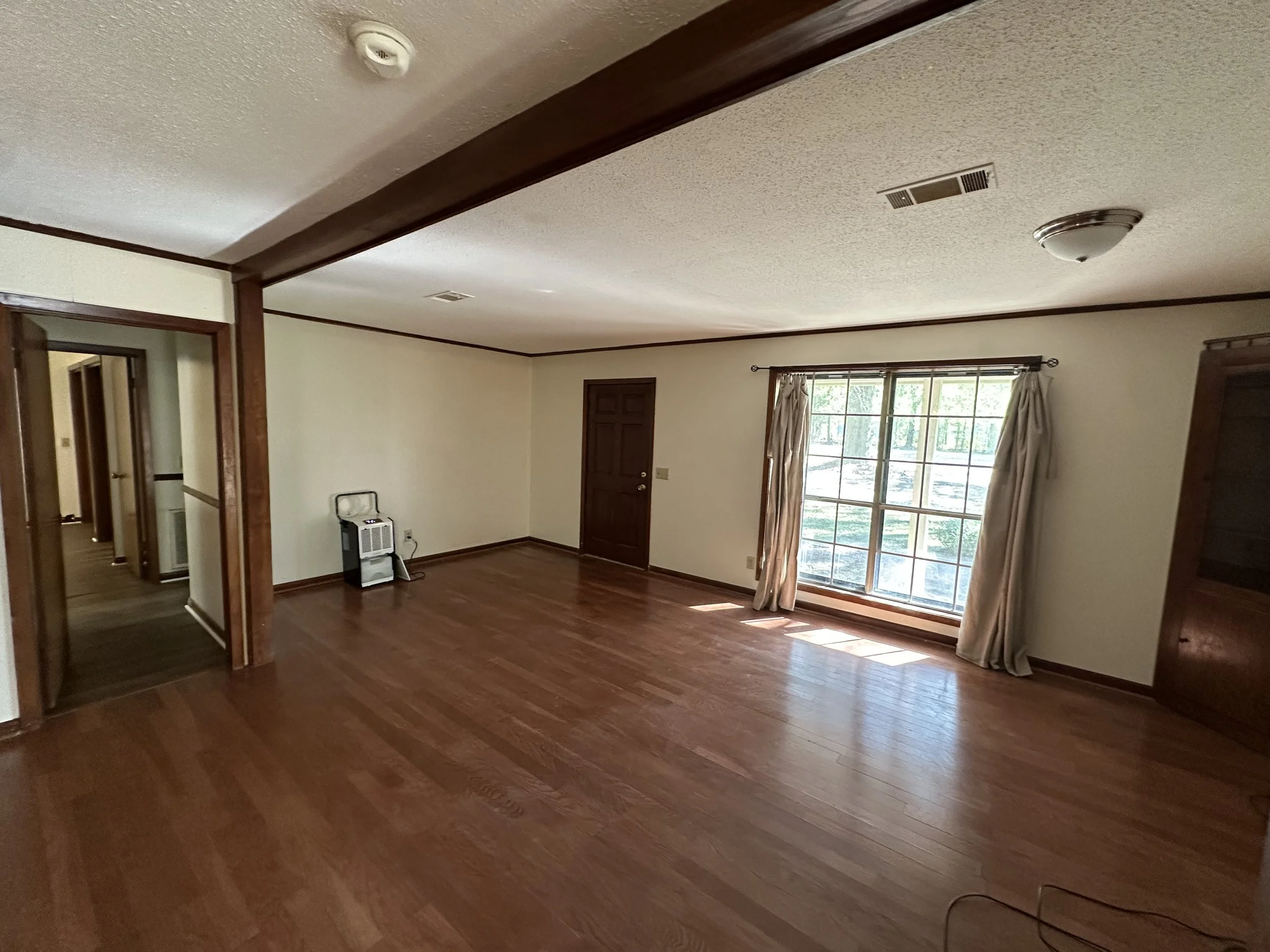 Empty living room with wooden floors, a large window with curtains, a ceiling light, and a small space heater against the wall.