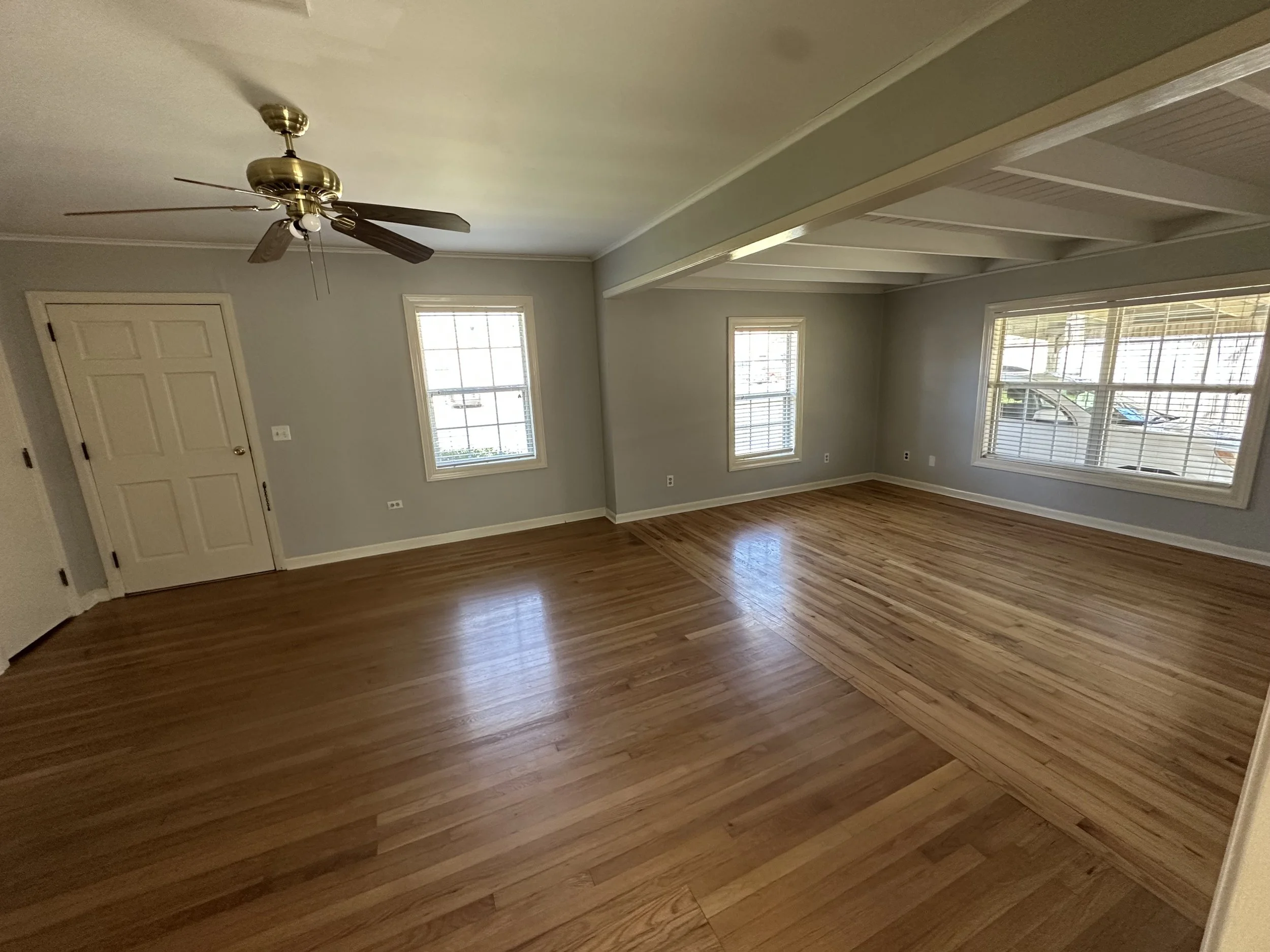 Empty living room with hardwood floors, light gray walls, three windows with white blinds, a white door, and a ceiling fan with brass accents and five blades.