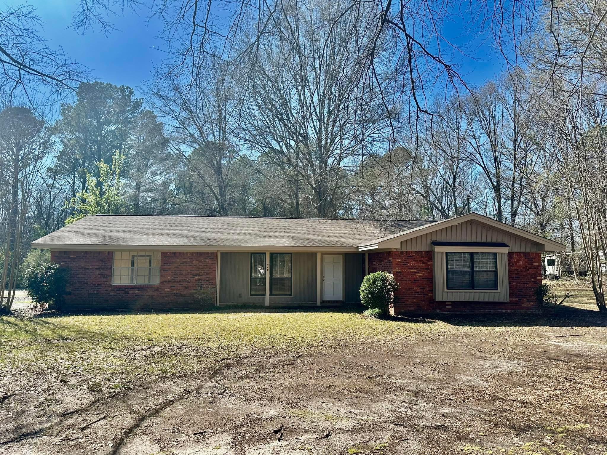 Single-story brick house with a gable roof, centered front door, and large windows, surrounded by trees and a gravel driveway.