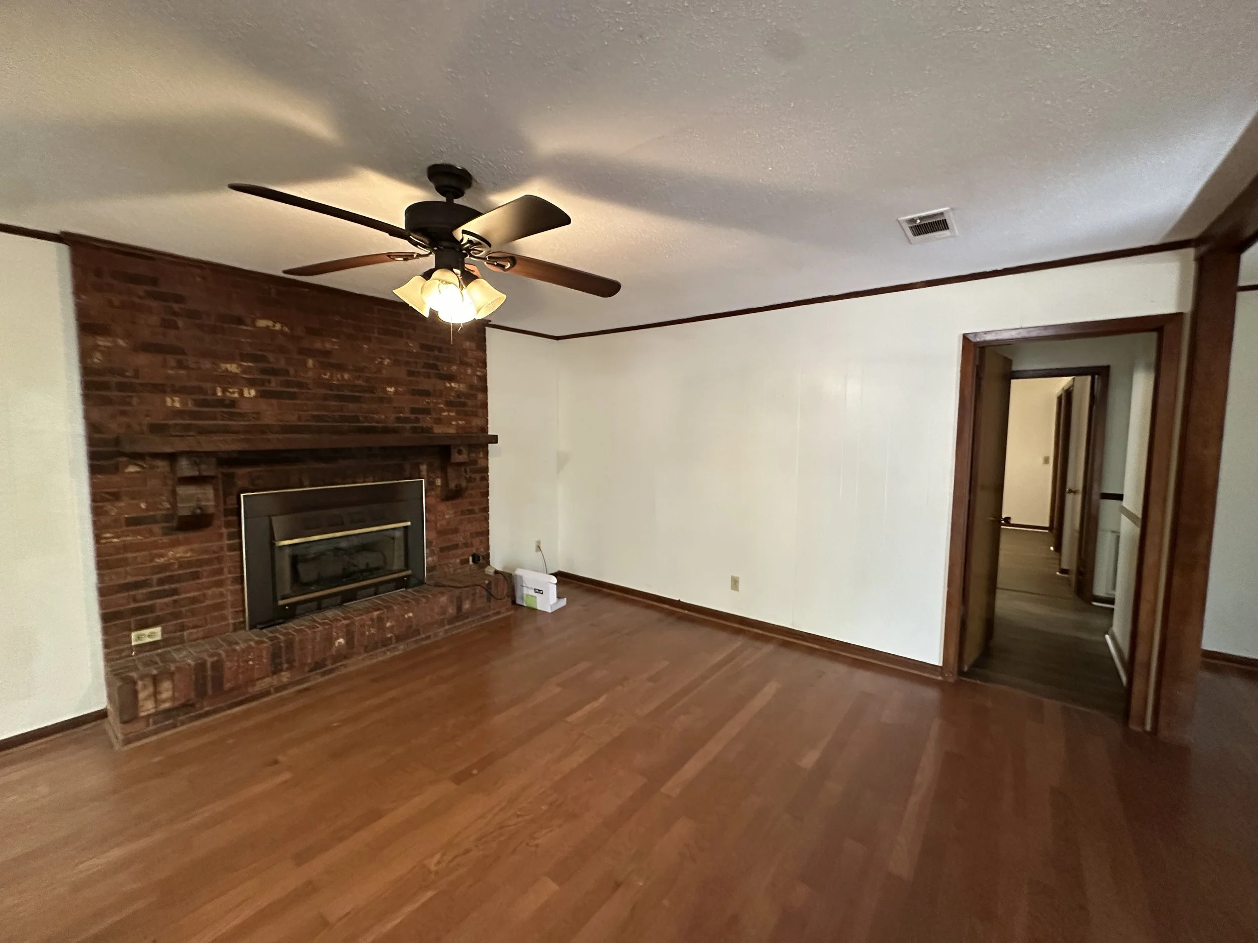 Living room with hardwood floors, brick fireplace, ceiling fan with light, and doorways leading to other rooms.