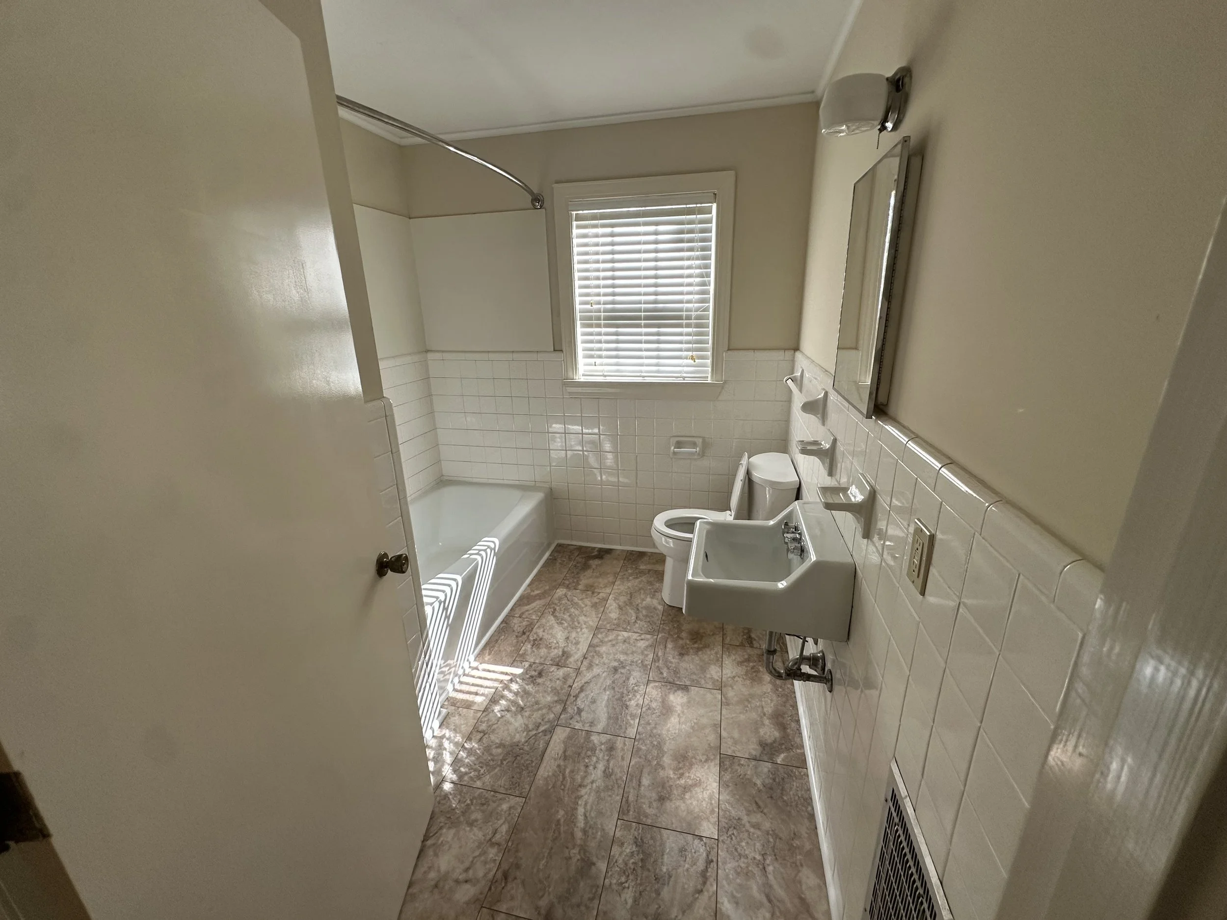 A bathroom with a bathtub, a toilet, a wall-mounted sink, a mirror, a window with blinds, and tiled walls and floor.