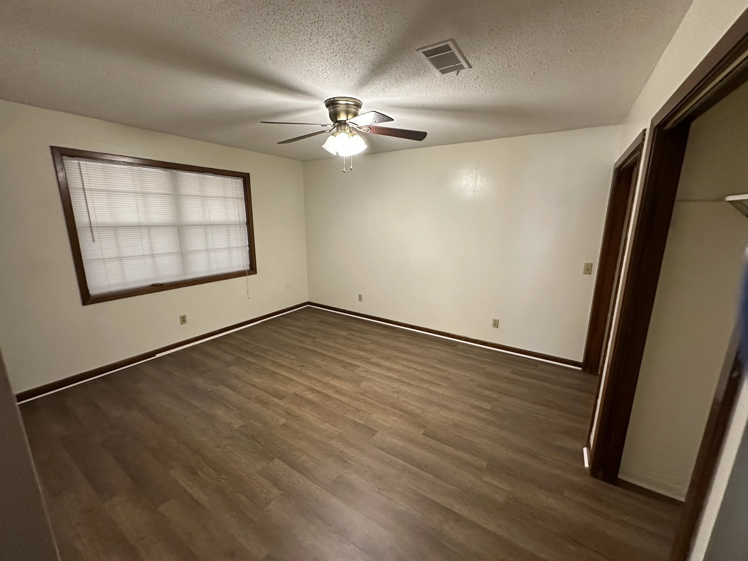 Empty room with wood flooring, white walls, a window with blinds, a ceiling fan with lights, and an open doorway leading to another room.