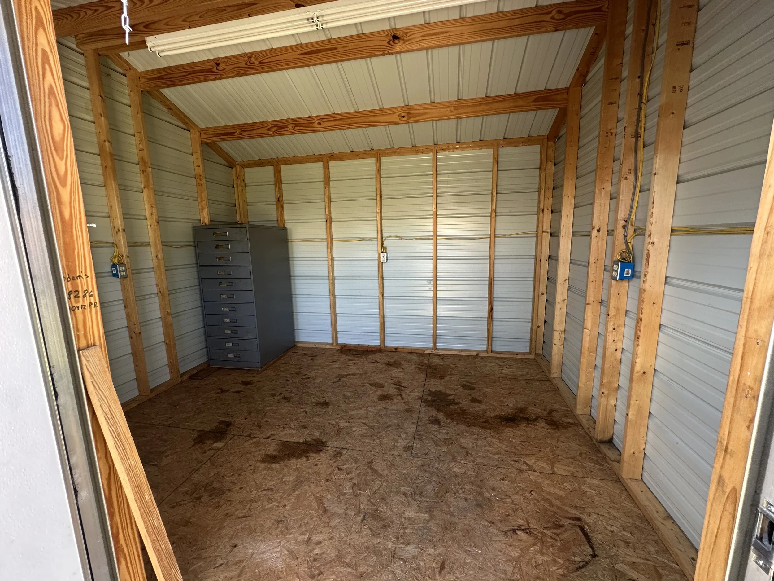 Empty storage unit with wooden framing, metal walls, and a gray filing cabinet in the corner.