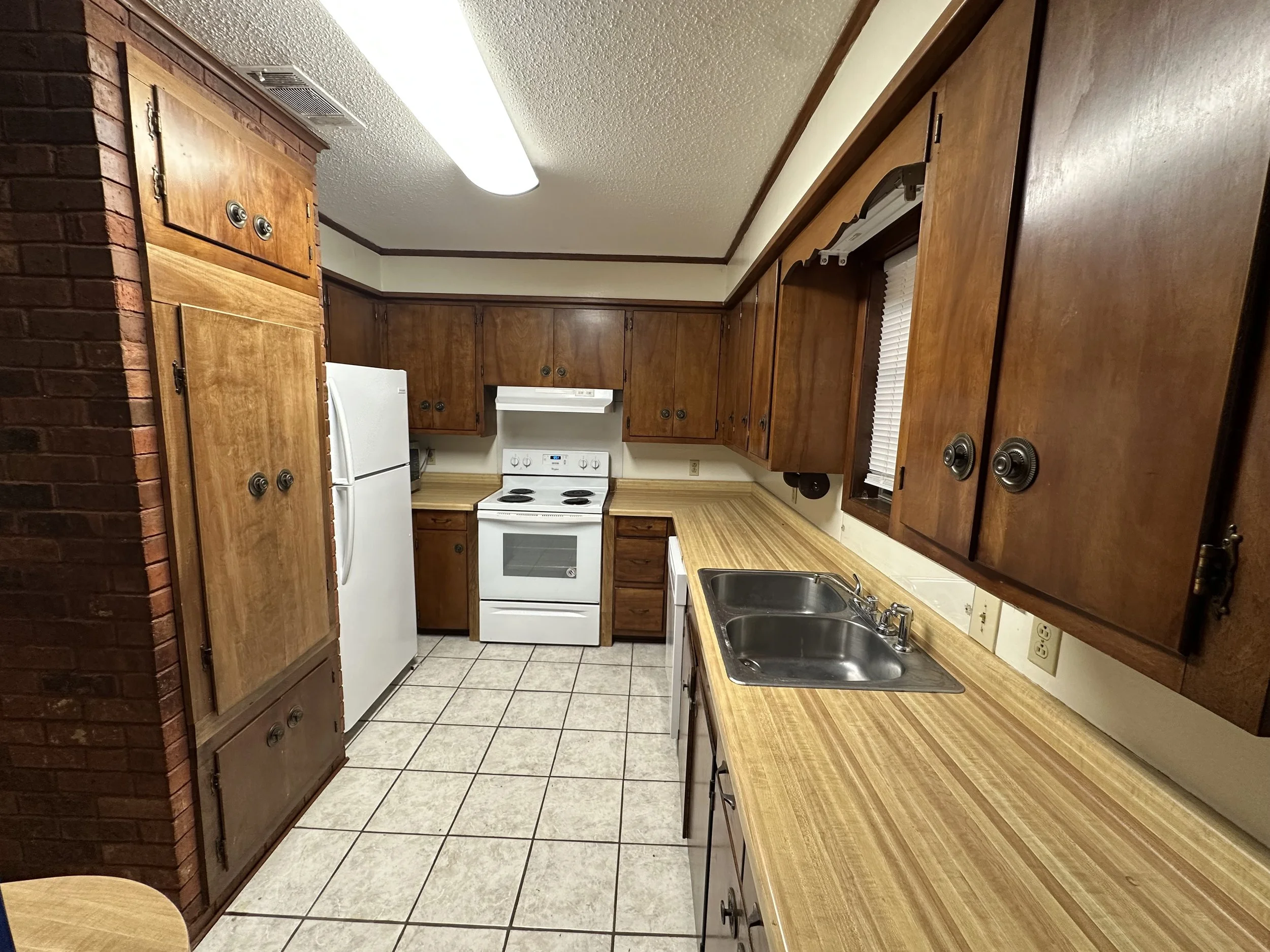 A kitchen with wooden cabinets, a white refrigerator, a white stove, a double sink, tiled floor, a window with blinds, and a fluorescent ceiling light.