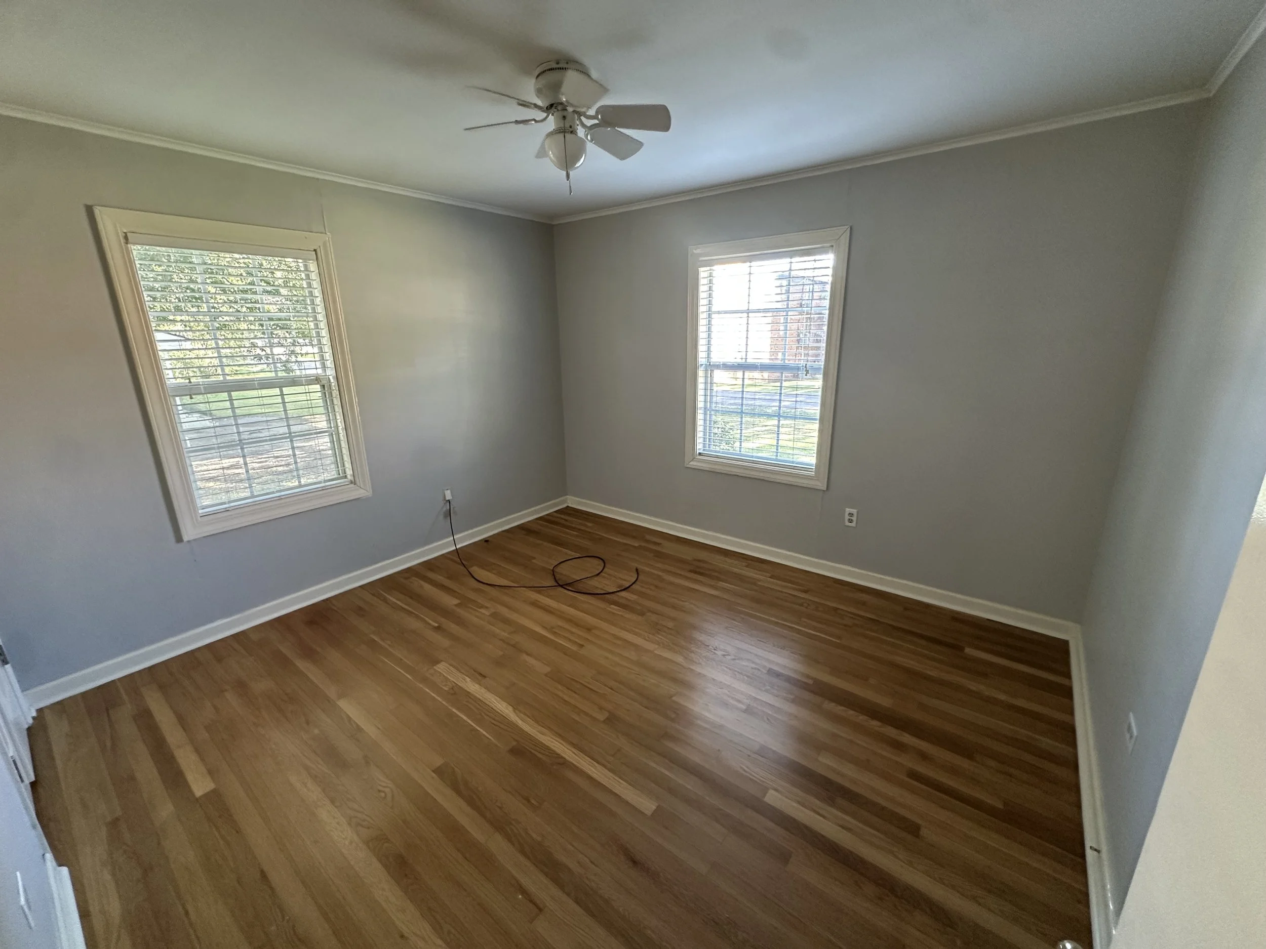 Empty room with hardwood floors, two windows with white blinds, gray walls, and a ceiling fan.