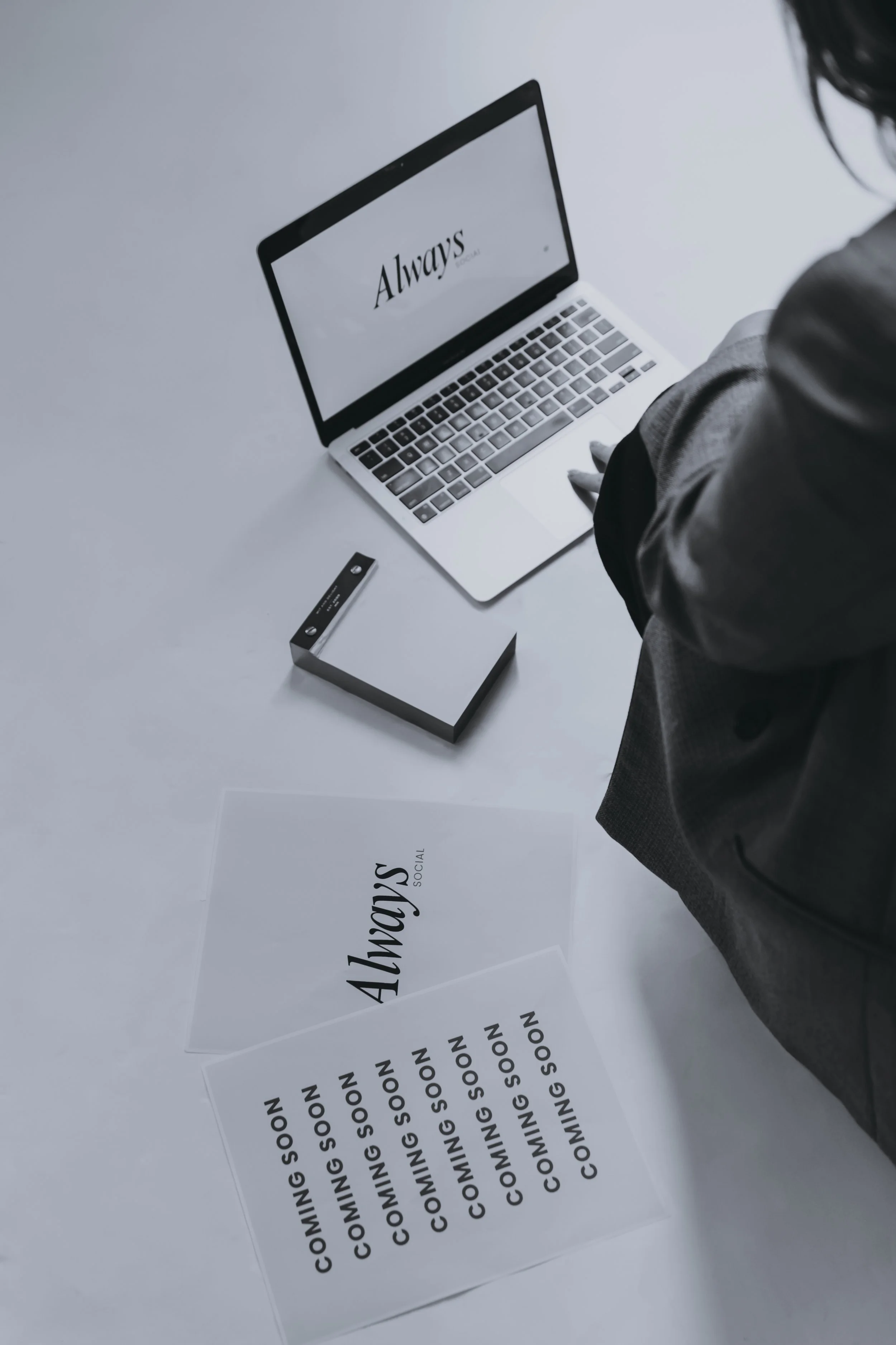 A person is sitting at a white table with a laptop displaying the word 'Always' on the screen, along with several signs that say 'COMING SOON' and one that says 'Always SOCIAL'.