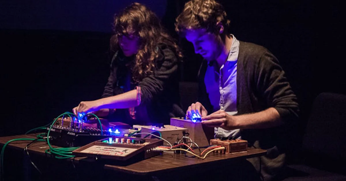 Two people working with electronic music equipment in a dark room, illuminated by blue and purple lights.