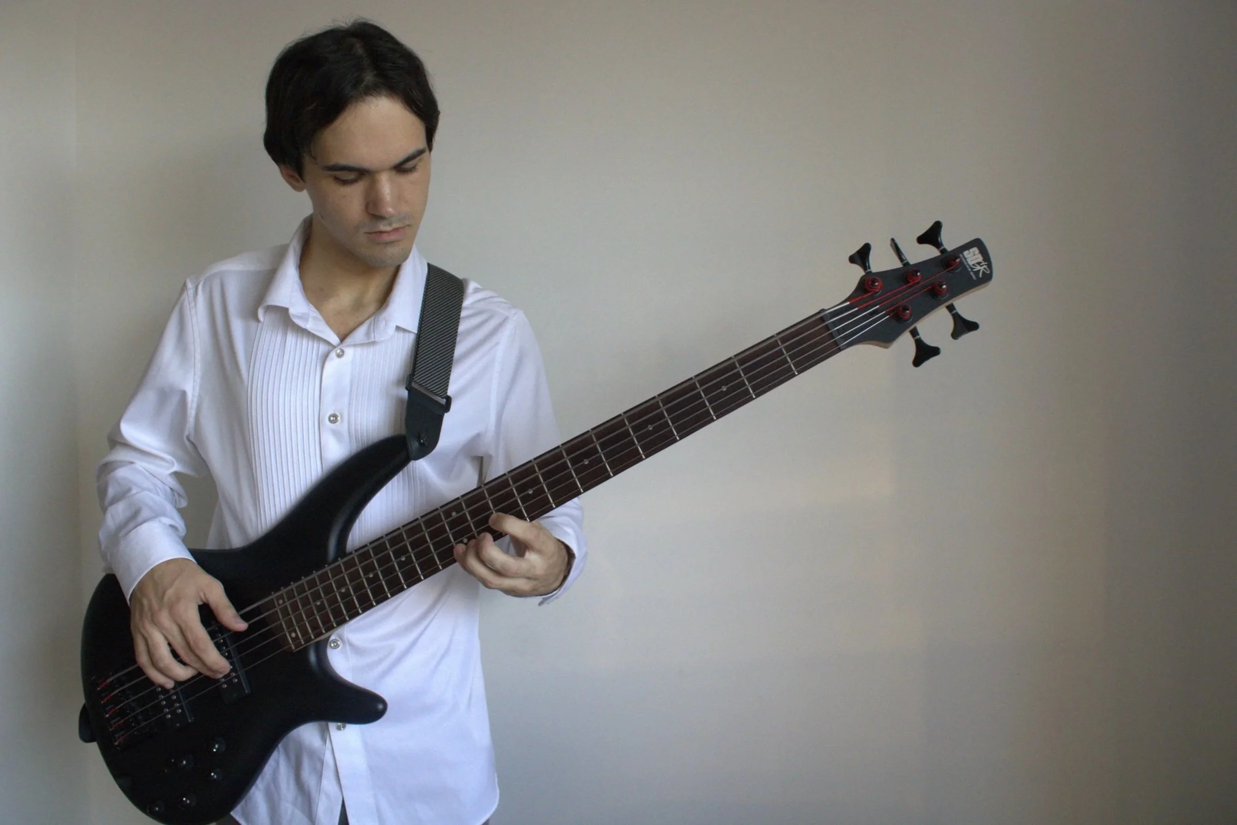 Chris is in a white shirt playing a black electric bass guitar against a plain light-colored wall.