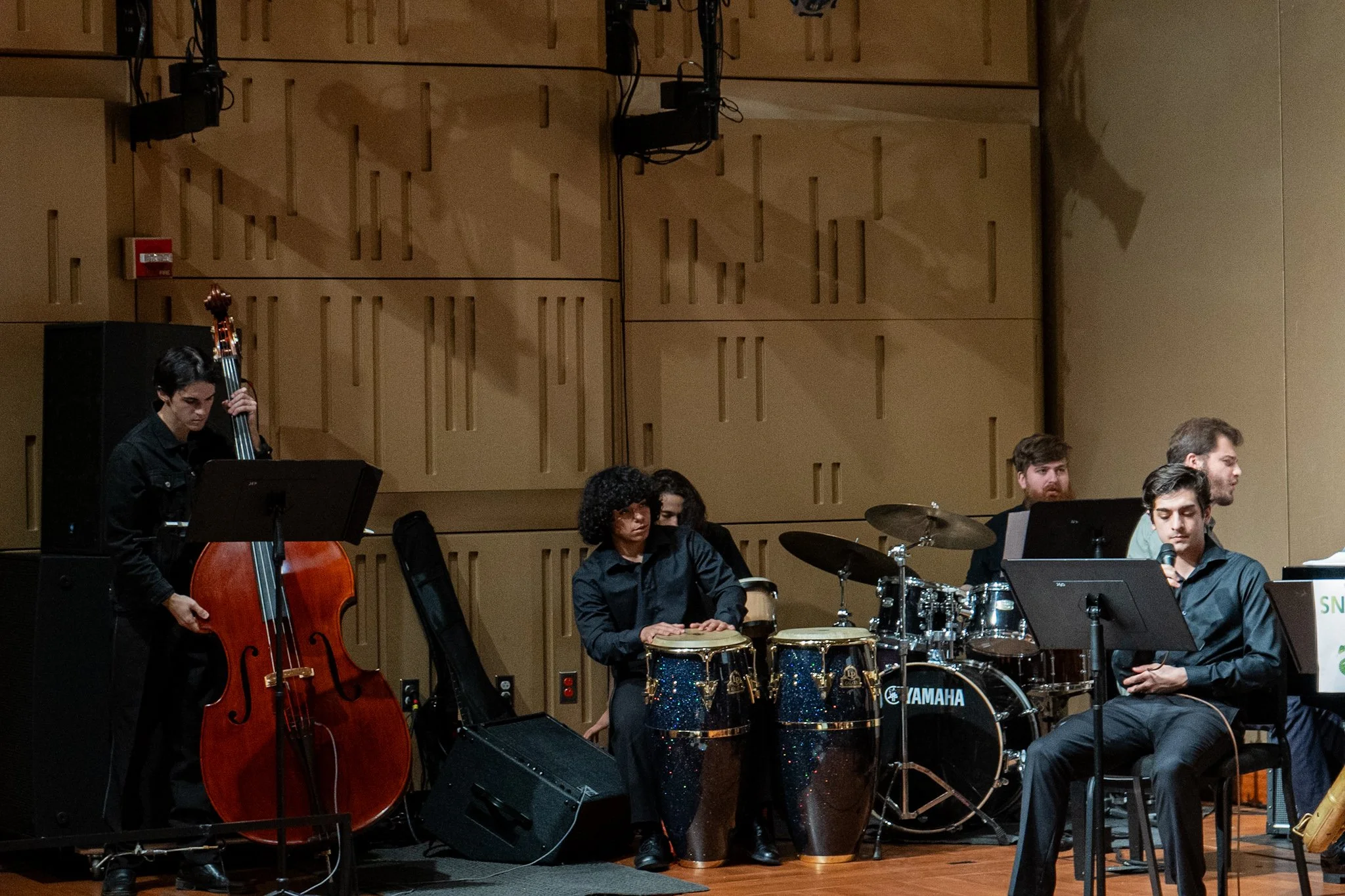 Jazz band performing on stage with musicians playing a double bass, conga drums, and other instruments, in a room with wooden acoustic panels.