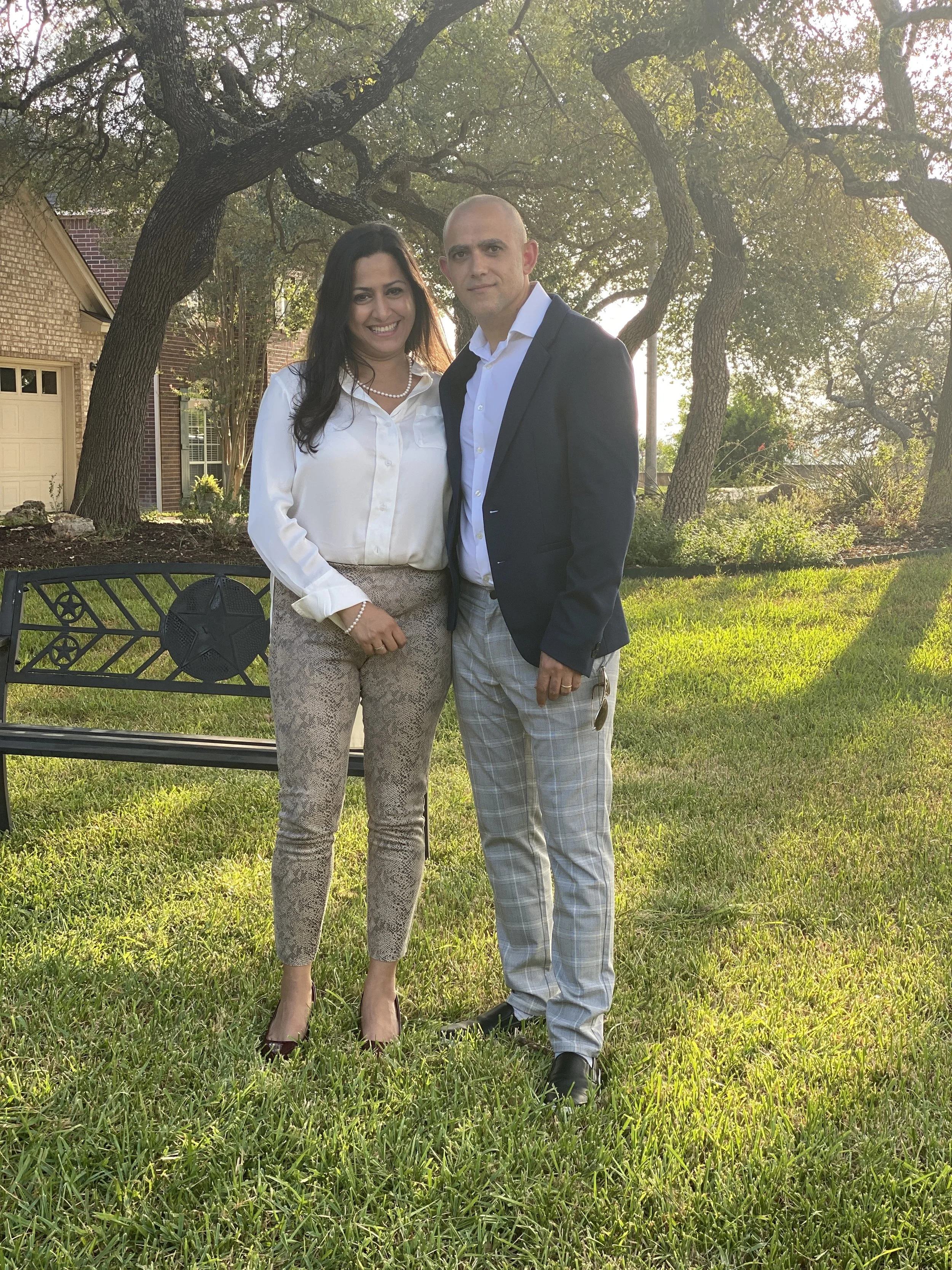 A man and woman standing together outdoors in a grassy yard, with trees and a house in the background, during sunset. The woman is wearing a white blouse, patterned pants, and high heels, while the man is dressed in a suit with plaid pants.