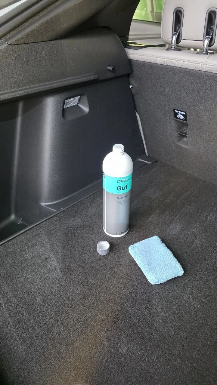 Open bottle of Gulf Gummint spray cleaner, blue sponge, and a small cap in the trunk of a car.