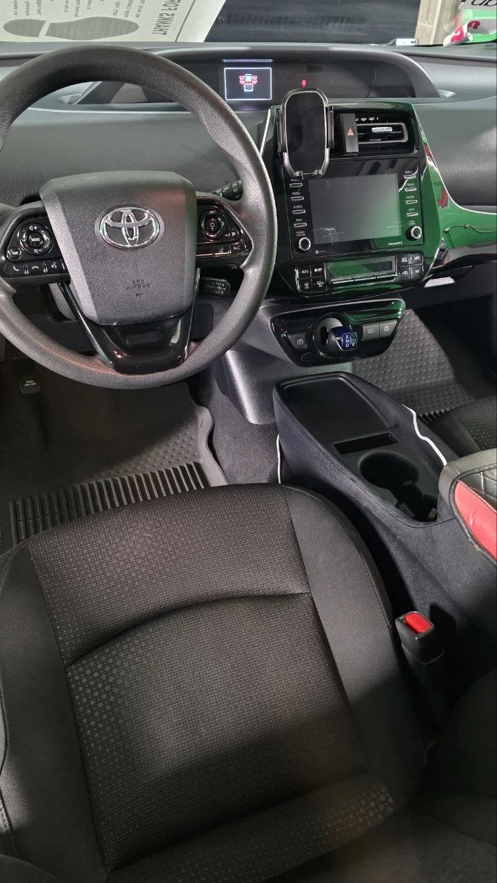 Interior of a Toyota vehicle's front cabin showing the steering wheel, dashboard with digital display, center console with touchscreen, and a black driver's seat.