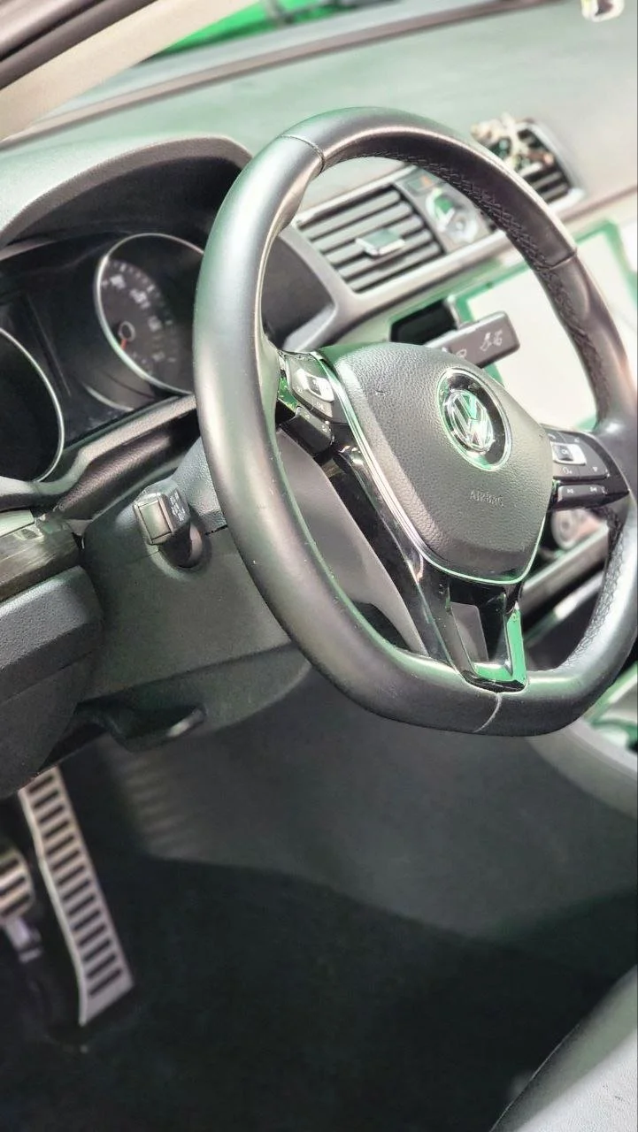 Interior of a Volkswagen car, focusing on the steering wheel with the Volkswagen logo, dashboard, and accelerator and brake pedals.