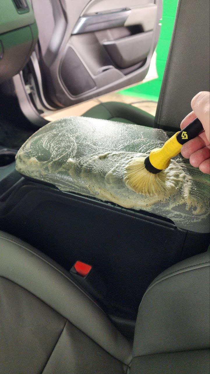 Someone cleaning car interior with a brush on a foam or sponge pad inside a vehicle.