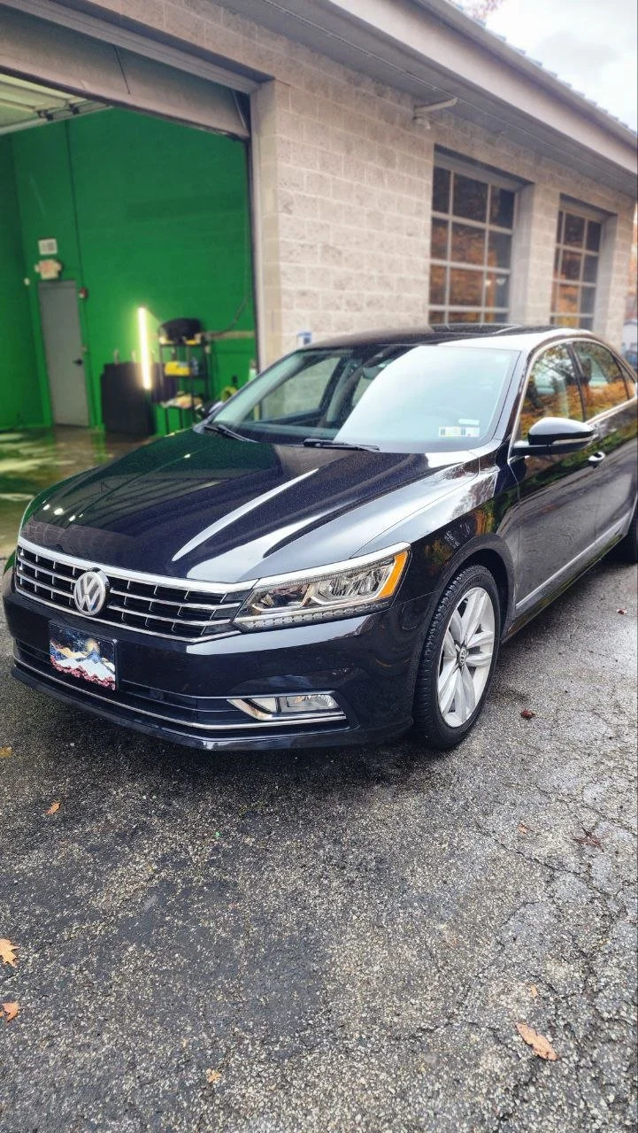 Black Volkswagen sedan parked outdoors near a brick building with large windows and a garage with green walls.