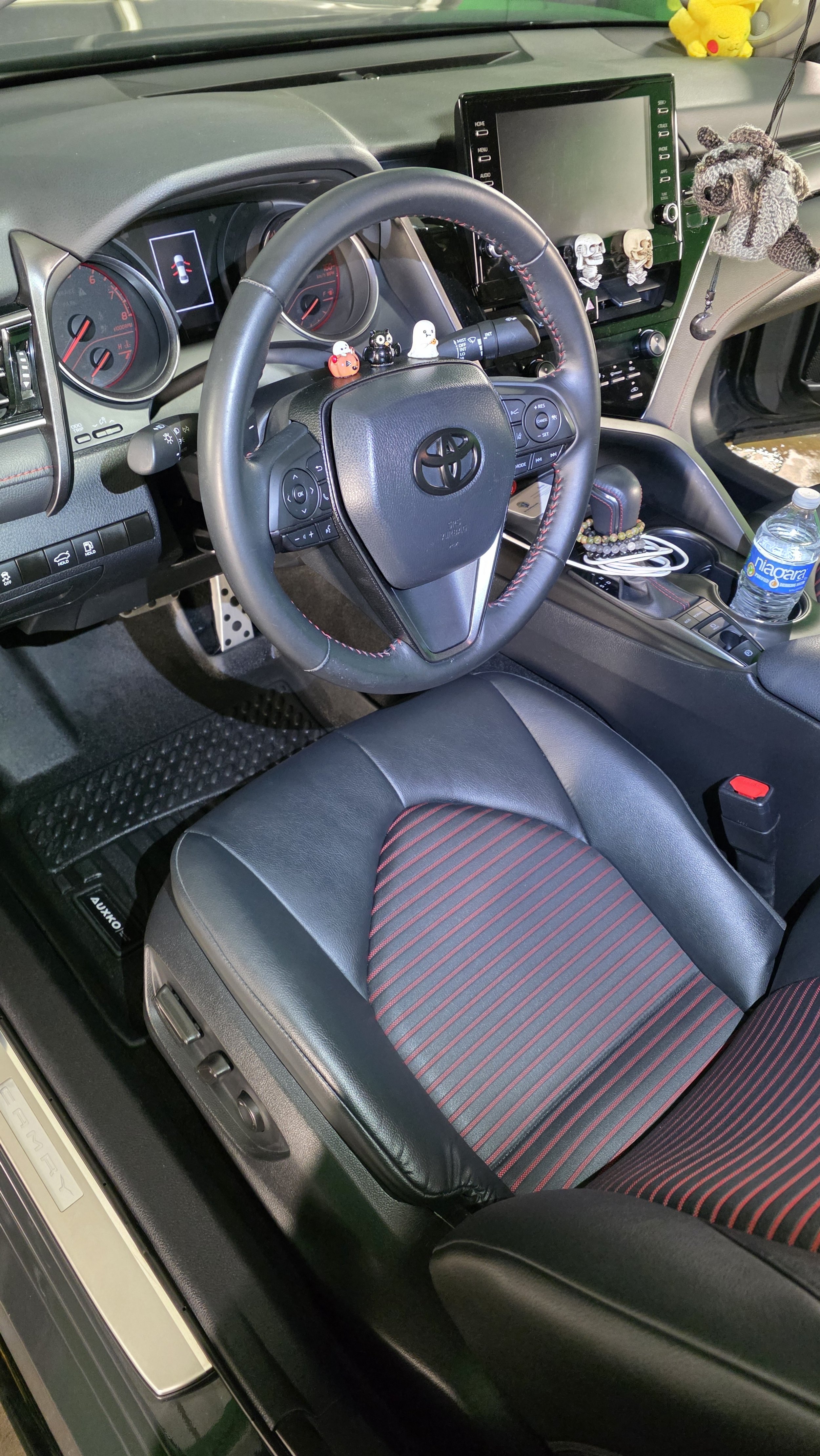 Close-up of the interior of a Toyota vehicle, showing the dashboard, steering wheel, and front passenger seat, with personal items like a water bottle, snacks, and plush toys visible.