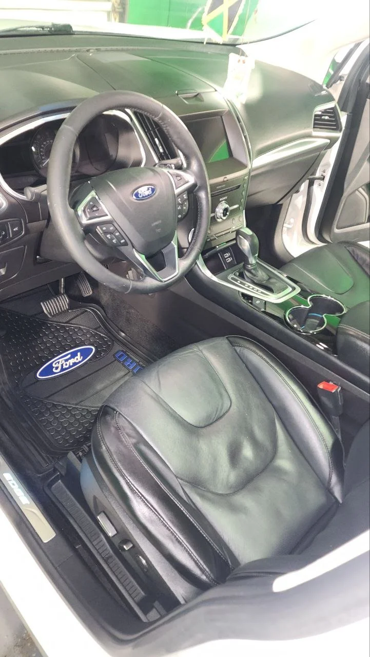 Interior of a Ford vehicle showing a leather seat, steering wheel with Ford logo, dashboard, gear shift, and floor mats with Ford logo.