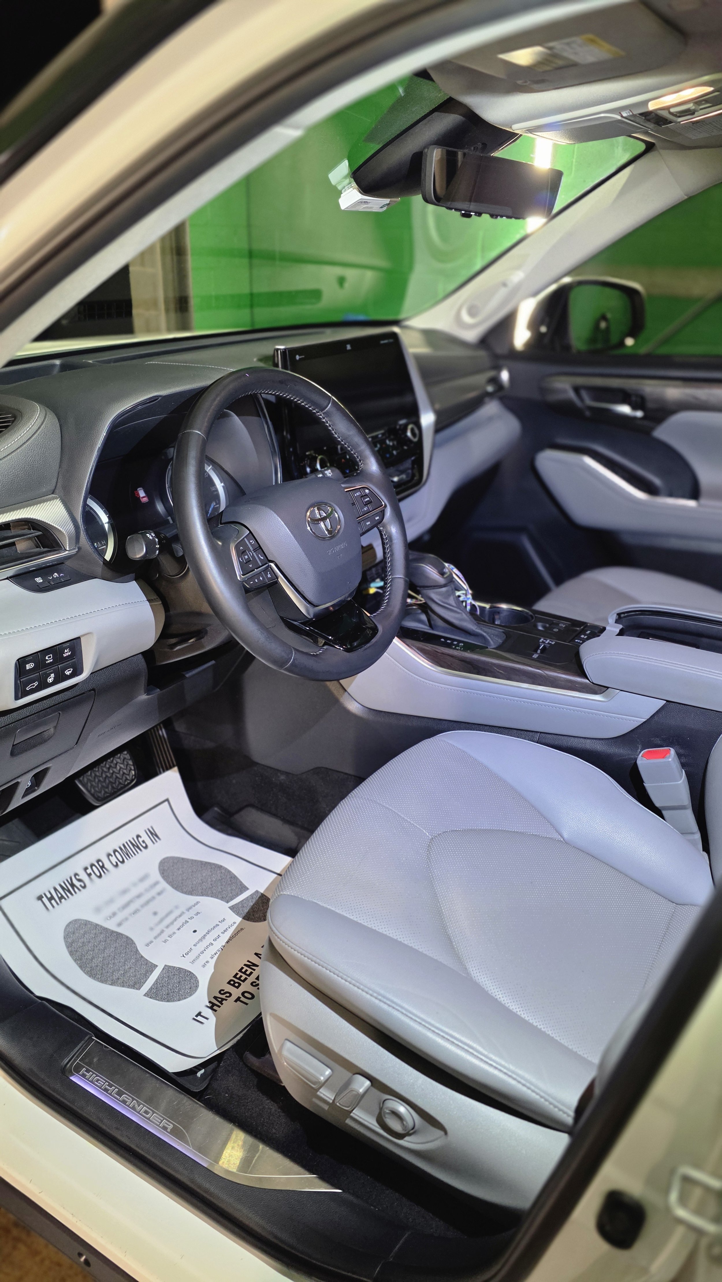 Interior of a Toyota Highlander showing the steering wheel, dashboard, front seats, and center console with gear shift. A paper floor mat with message for customers is on the driver's side floor.