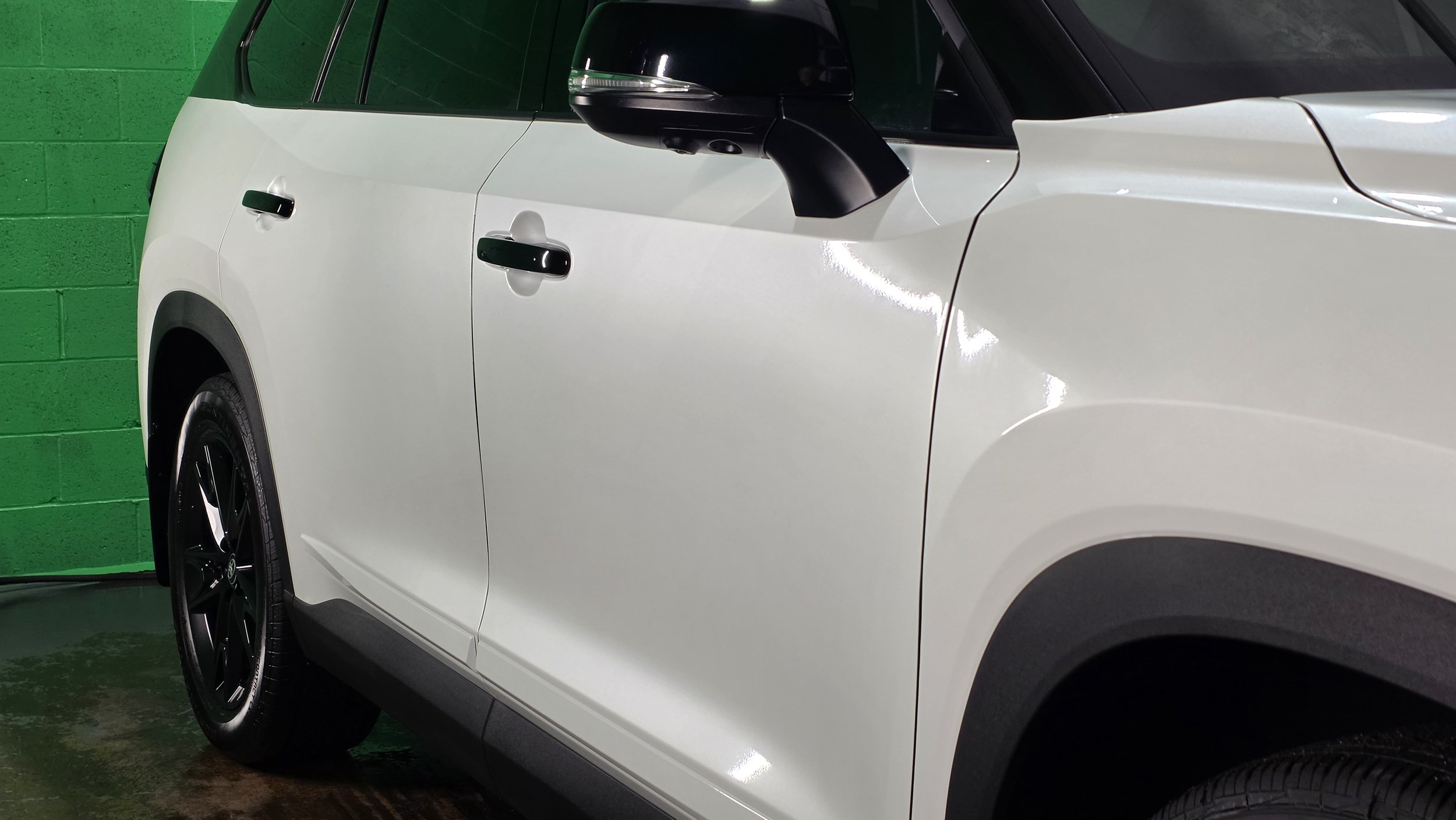 Close-up of a white SUV with black side mirrors and door handles, against a green wall background.