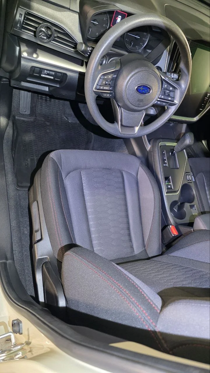 The image shows the interior of a Subaru vehicle, focusing on the driver's seat, steering wheel, and dashboard. The steering wheel has controls on both sides, and there is an infotainment screen to the right of the steering wheel. The gear shifter is