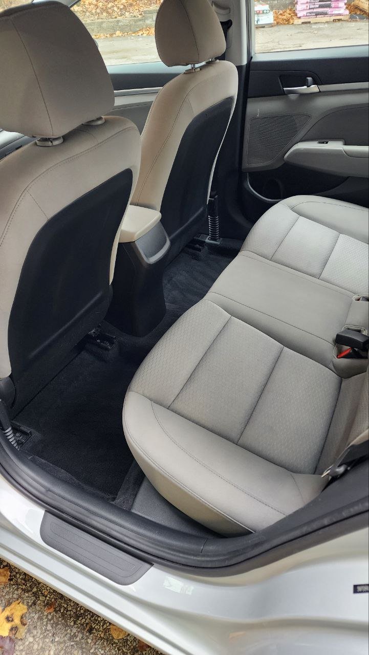 Interior of a car showing the back seat area with light gray upholstery and the front seats with beige and black upholstery. The car door is open, and part of the ground with some leaves is visible outside.