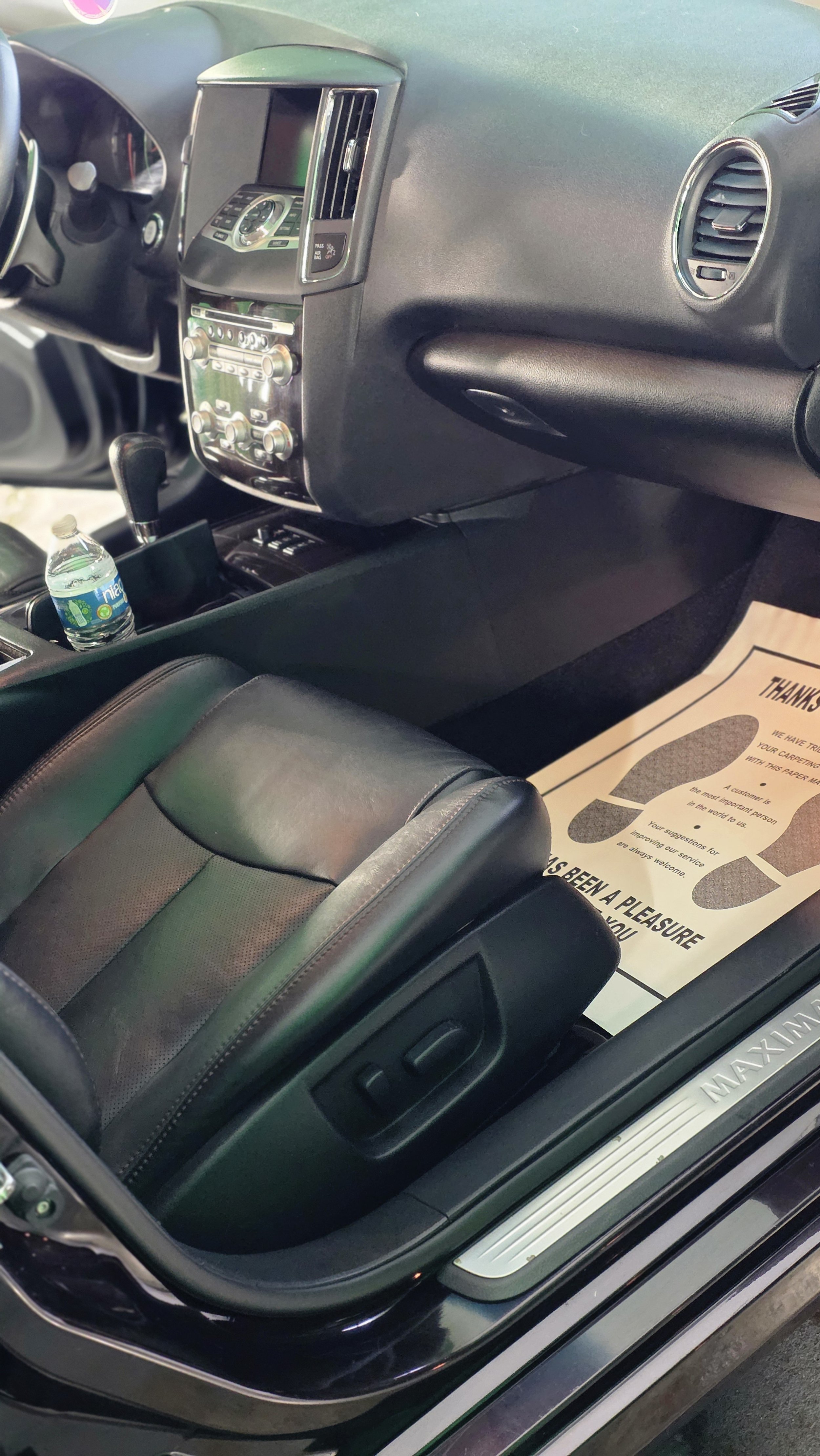 The interior of a car shows the front passenger seat, center console with a water bottle, dashboard with controls, and a floor mat with printed text.