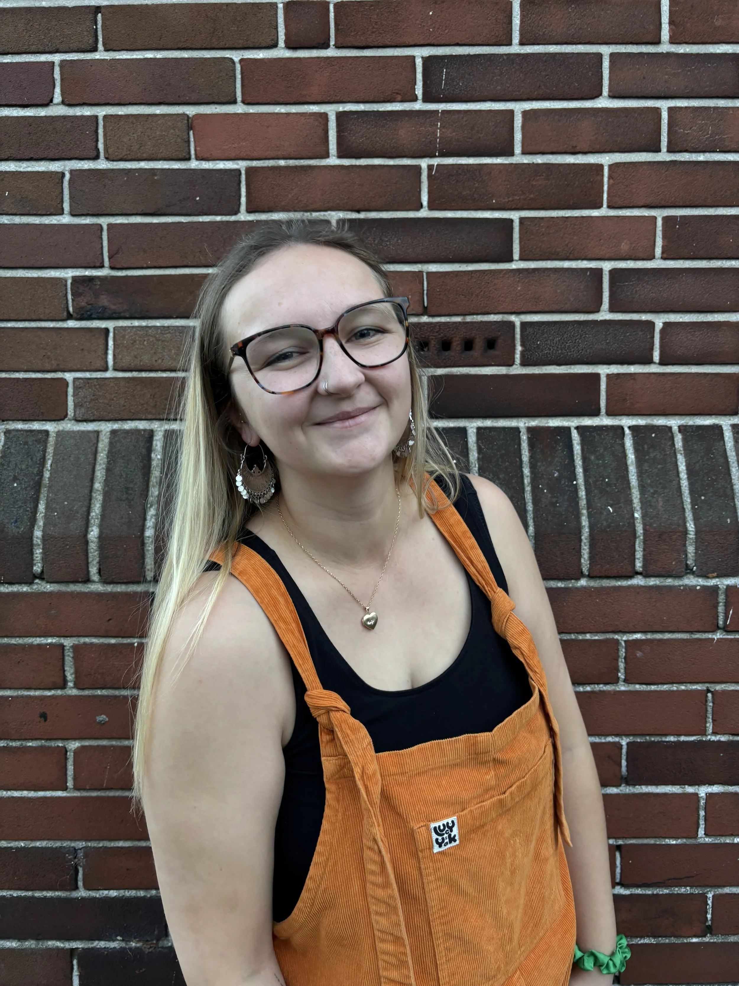 A woman with blonde hair, glasses, and a nose piercing, smiling, stands in front of a brick wall. She is wearing a black top, orange overalls, earrings, a gold heart necklace, and a green bracelet.