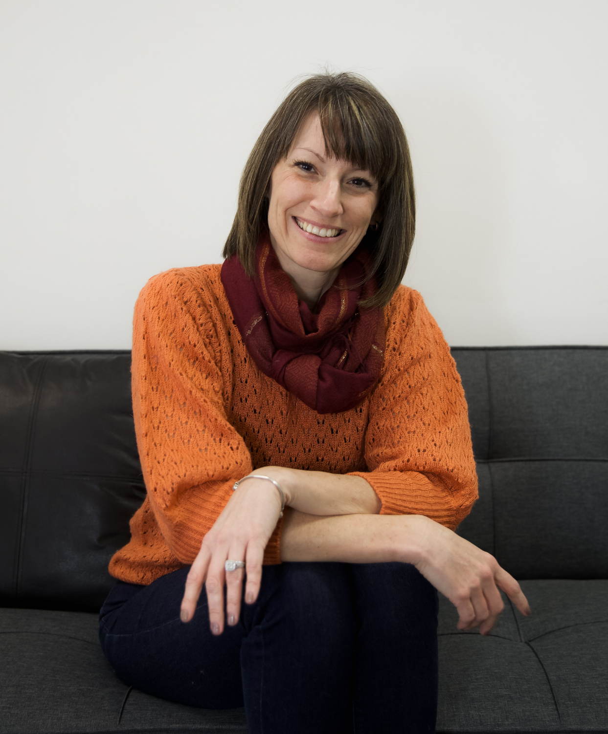 A woman with brown hair, wearing an orange sweater and a red scarf, sitting on a black couch against a plain white wall, smiling at the camera.