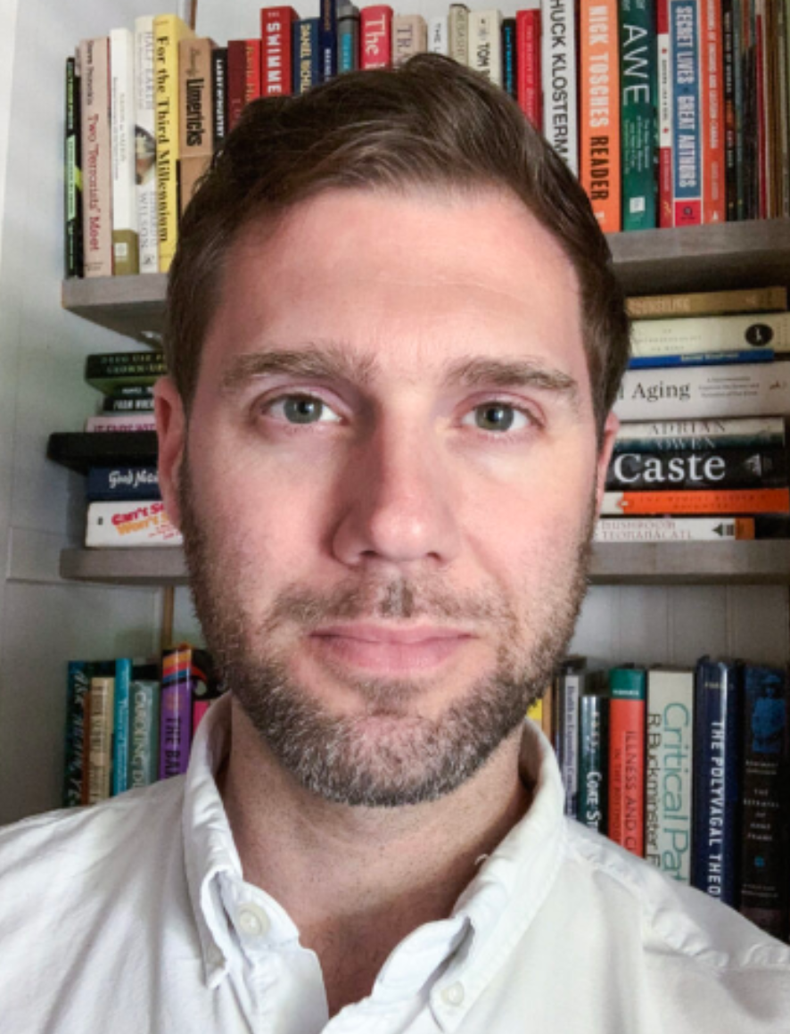 A man with brown hair and blue eyes taking a selfie in front of a bookshelf filled with various books.