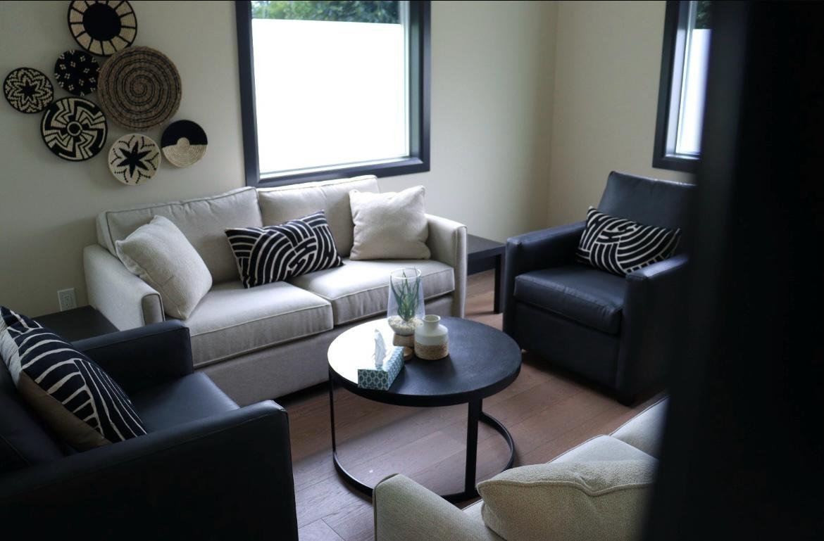 Living room with a cream sofa, black armchairs, wooden flooring, a black circular coffee table with decorative items, multiple black and white pillows, and wall art with woven baskets.