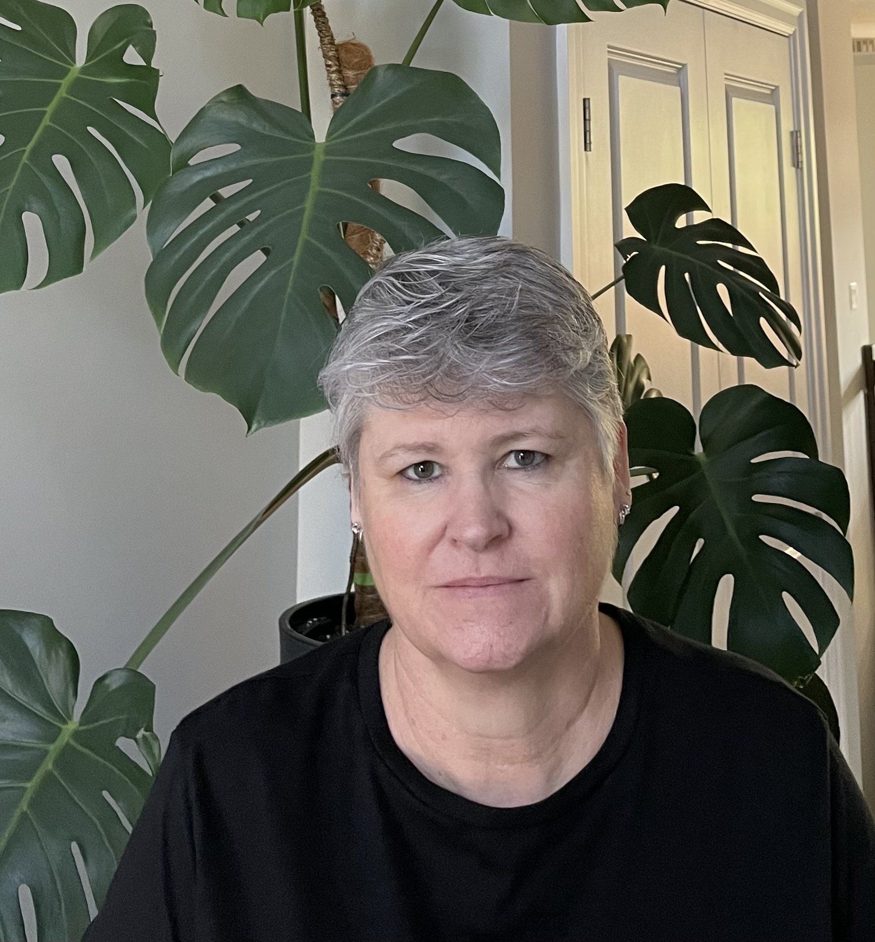 A woman with short gray hair wearing a black shirt standing in front of green Monstera plant leaves.