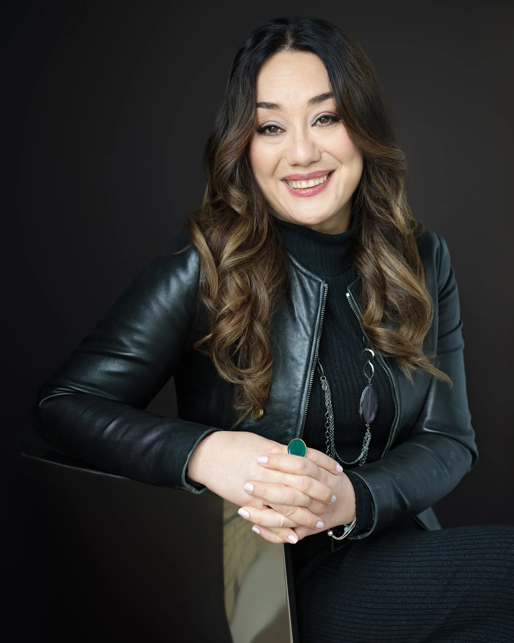 A woman with long wavy brown hair smiling and wearing a black leather jacket, black top, and jewelry, posed against a dark background.