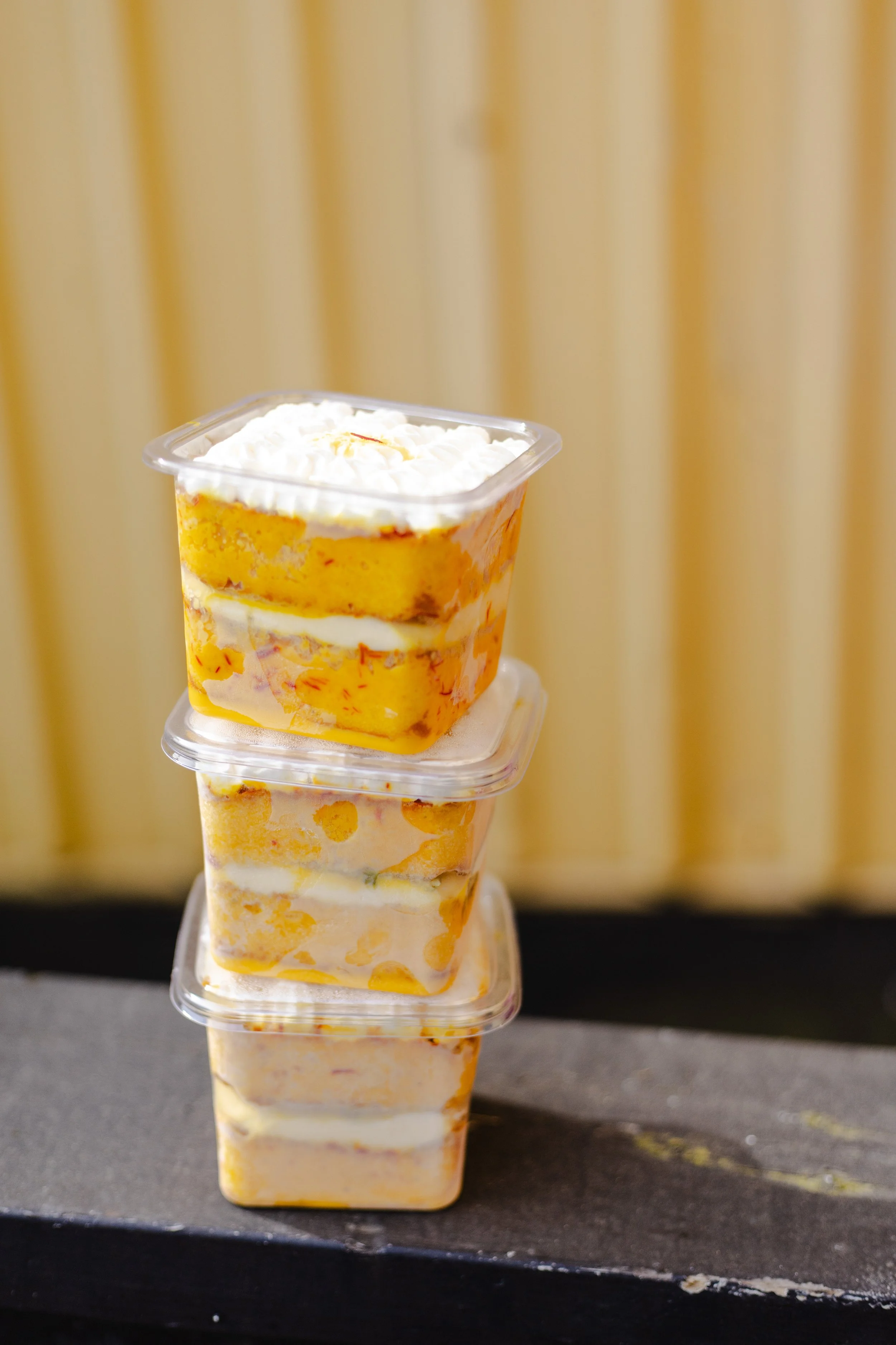 Stack of four clear plastic containers filled with Rasmalai Cake topped with whipped cream, placed on a dark surface with a light-colored striped background.