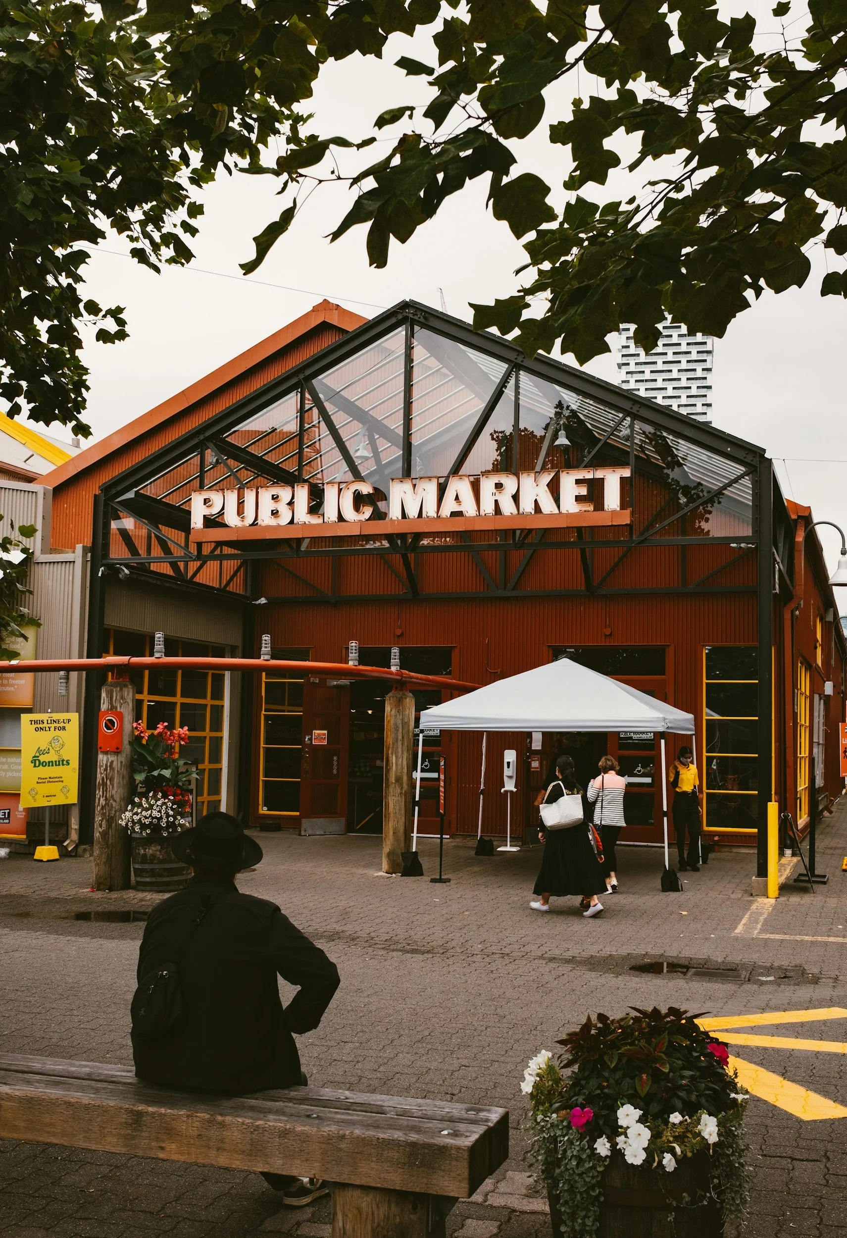 People entering and waiting outside a red building with a sign that reads 'Public Market.' Sitting on a wooden bench in the foreground, a person in black with a hat and backpack. There is a white canopy and a flower pot with colorful flowers.