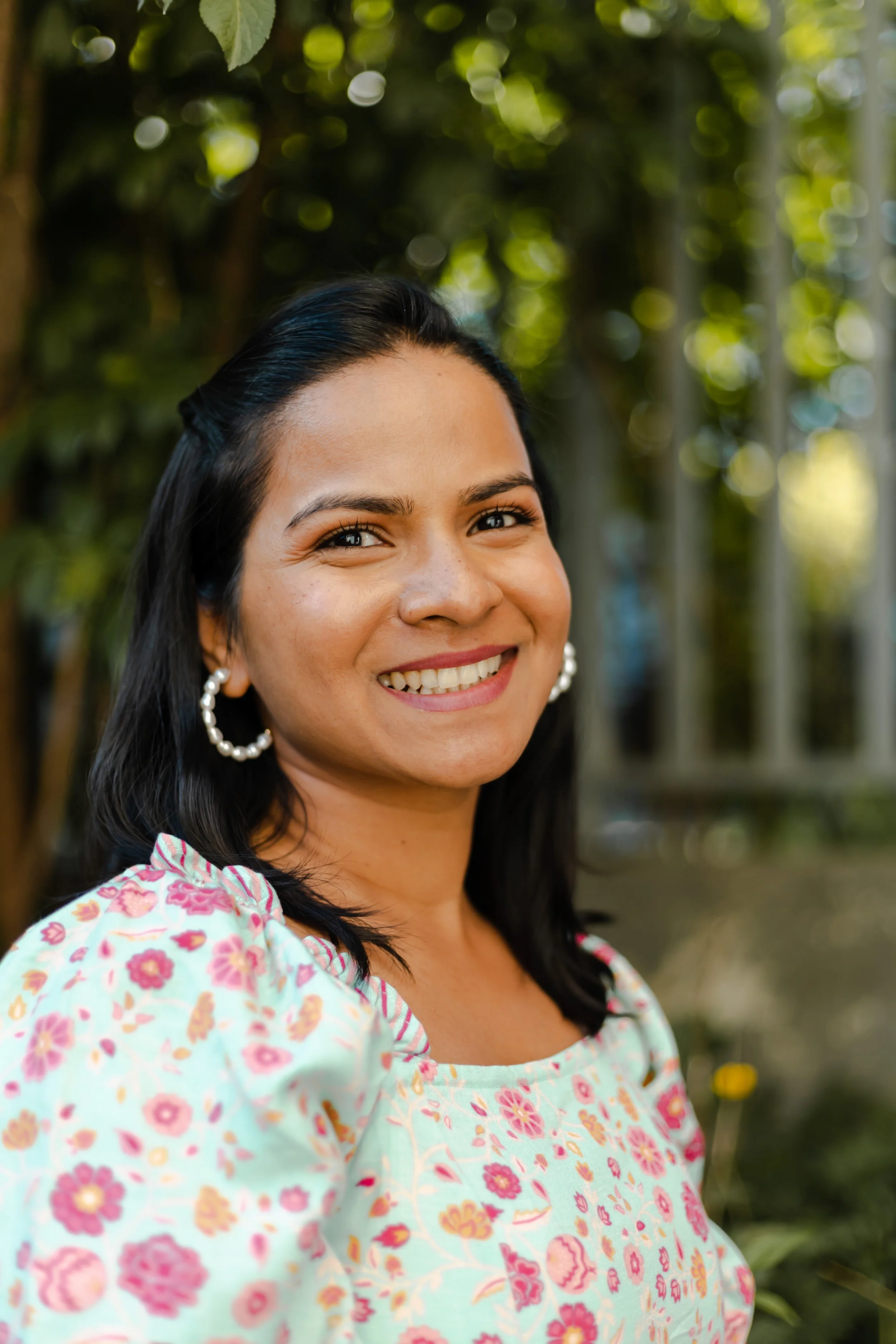 Smiling woman with dark hair, wearing pearl earrings and a floral dress, outdoors with green trees and sunlight in the background. Woman in photo is owner and baker of Elaichi Patisserie, Vinaya.